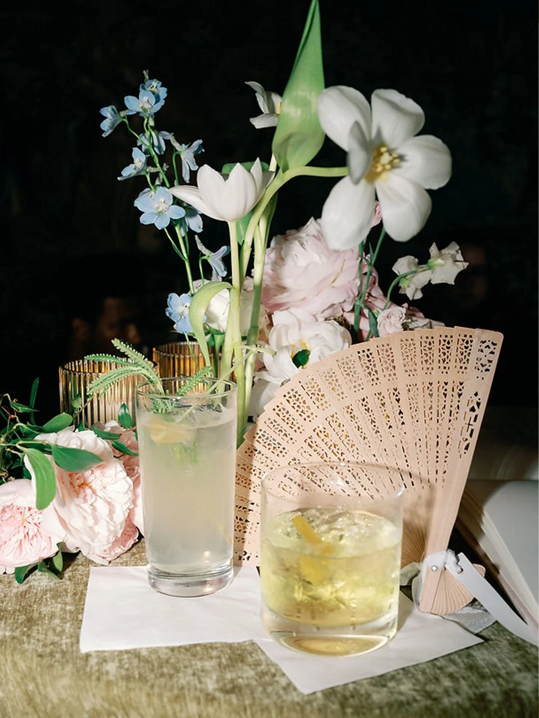 A table with two beverages, one in a tall glass with a lemon slice and mint leaves, and another in a short glass with a lemon slice, surrounded by pink and white flowers, a decorative fan, and two gold candle holders.
