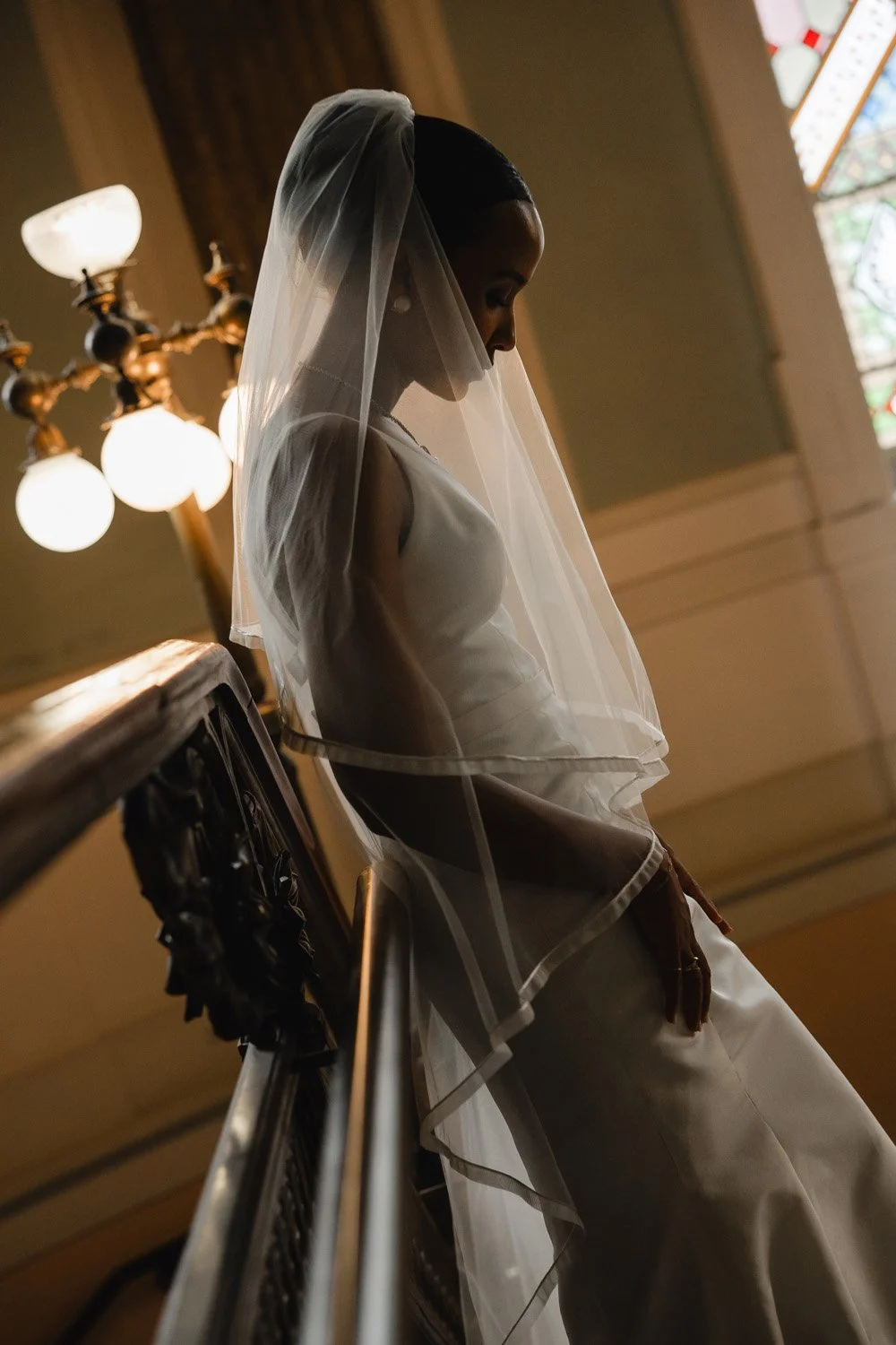 A bride dressed in white with a veil standing indoors near a staircase, with warm lighting from a chandelier and stained glass windows in the background.