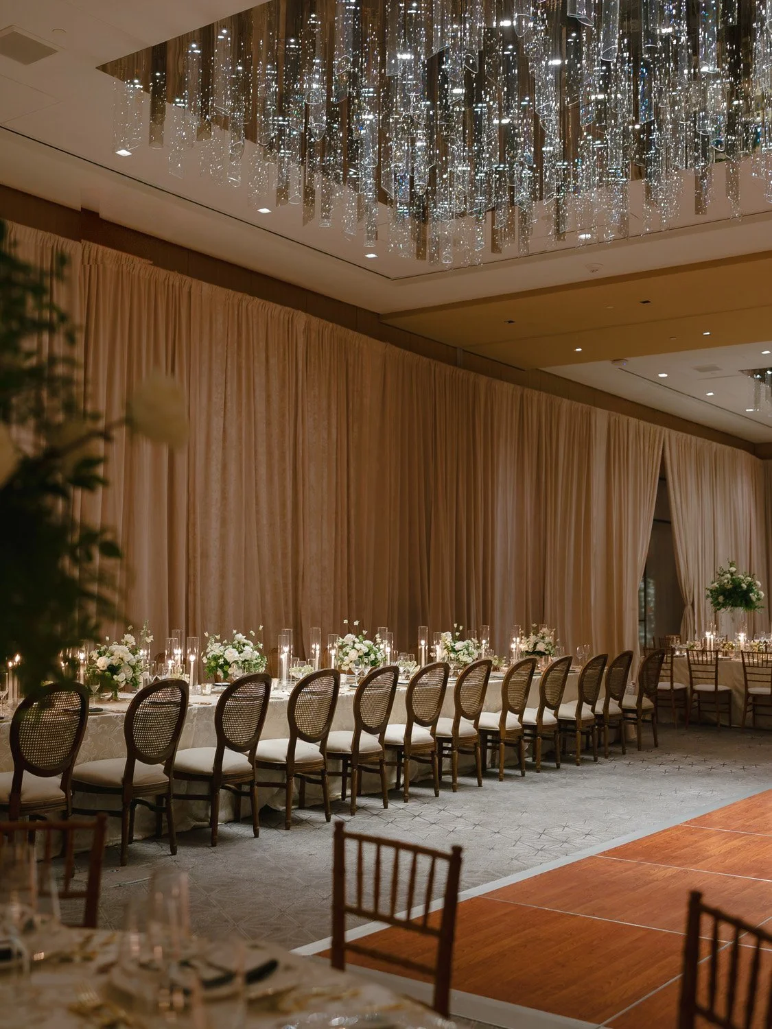Elegant banquet hall with a long table decorated with white flowers and candles, beige drapes, and a modern chandelier with hanging glass accents.