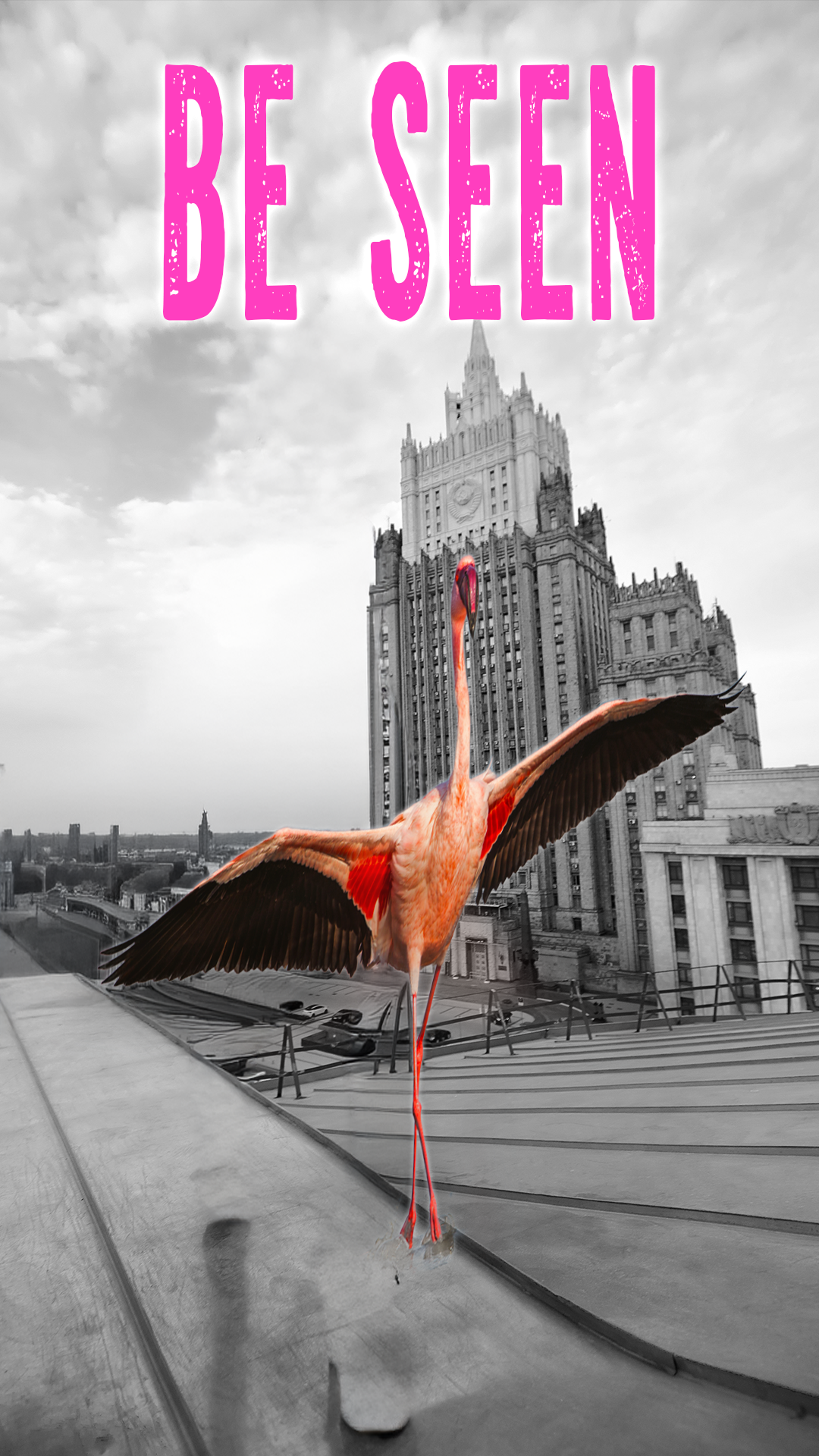A pink flamingo with its wings spread standing on a rooftop ledge in front of a tall, gothic-style building. The background is black and white, and the words "BE SEEN" are written at the top in large, pink, distressed letters.