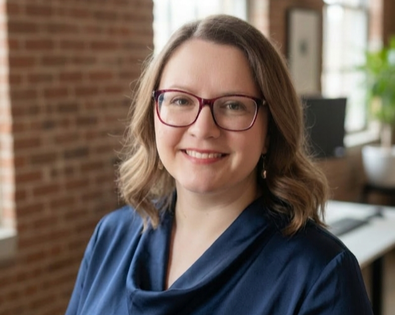 A woman with shoulder-length wavy hair and glasses smiling in an indoor setting with exposed brick wall and a computer in the background. She is the founder of Water's Edge Poetry, Erin Rebant.