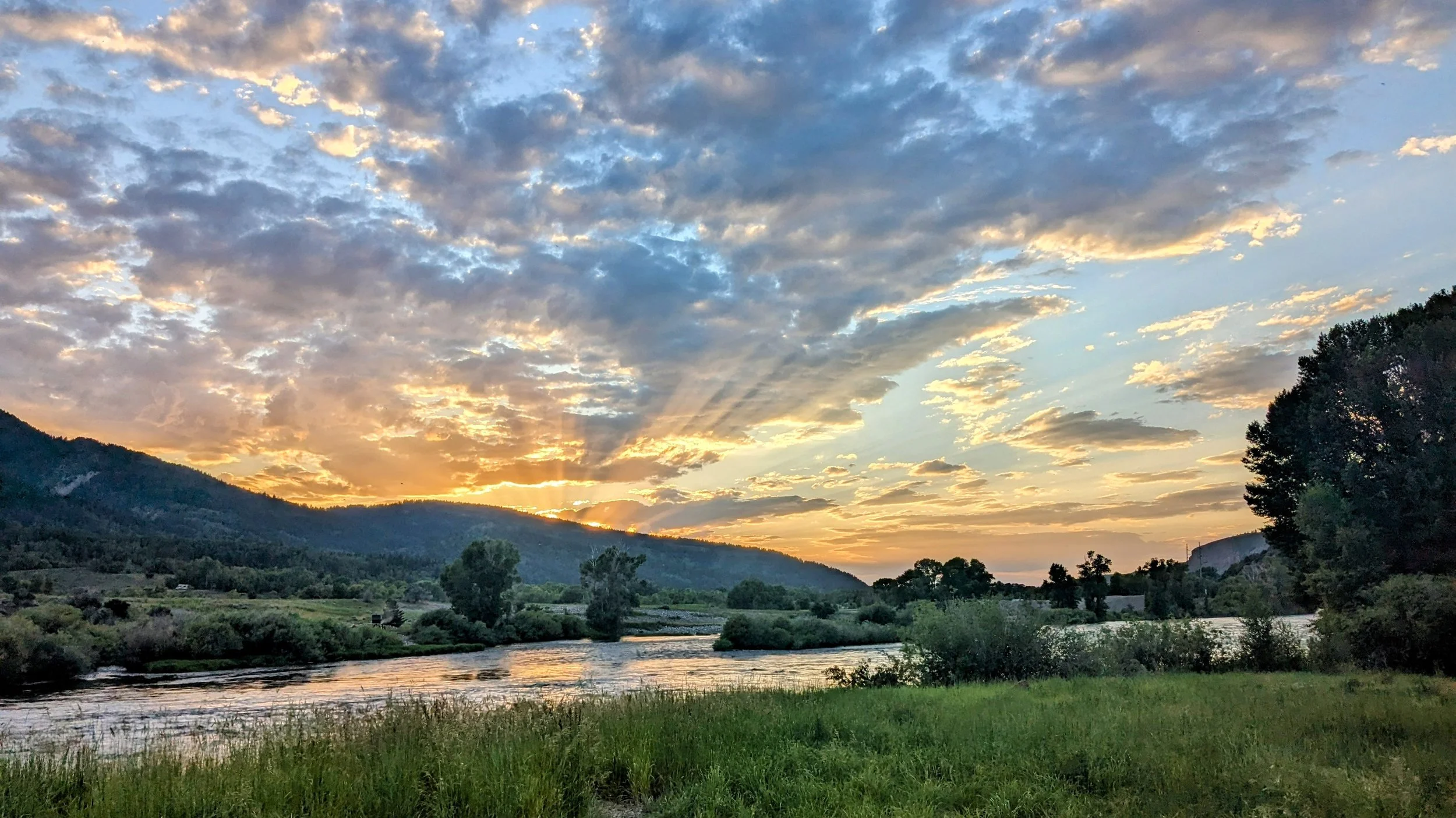 Sunset over a river with green grass in the foreground, trees along the riverbank, hills in the background, and rays of sunlight breaking through the clouds.