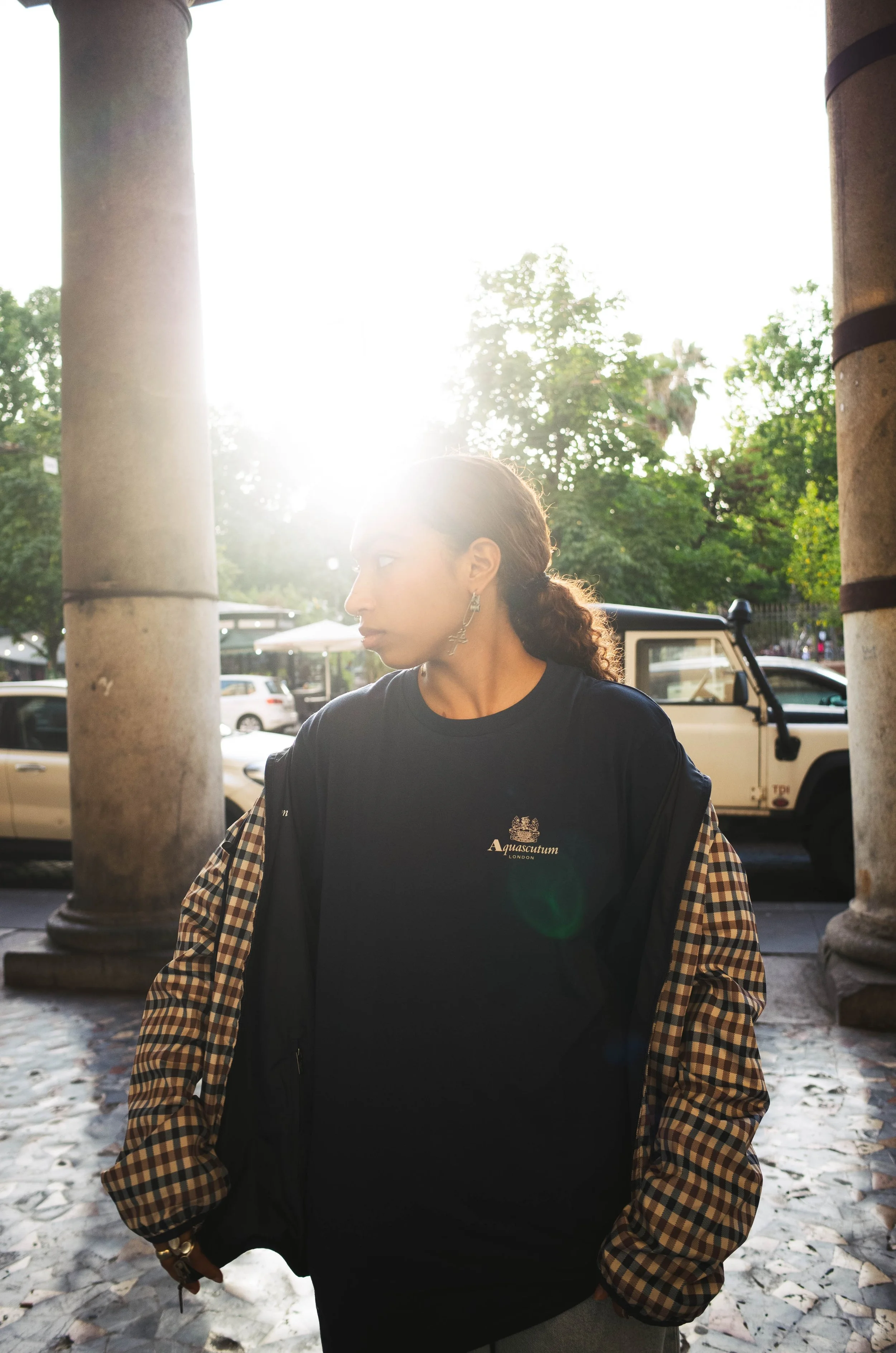 A woman with curly hair wearing earrings and a black shirt with the logo 'Aquascutum London' and a checkered jacket, standing outdoors near large stone columns with parked cars and trees in the background, backlit by the sun.