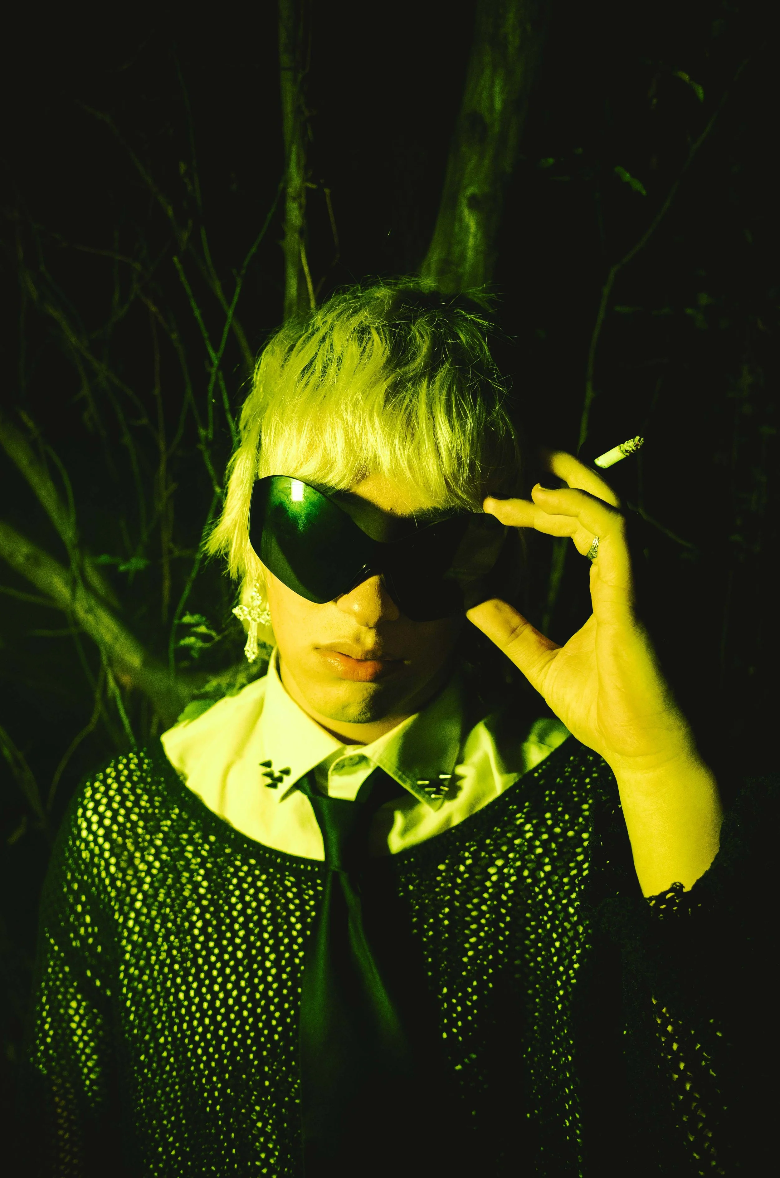 A person with short, blond hair wearing dark sunglasses, a white shirt with black stars on the collar, a black tie, and a black sequined jacket. They are holding a cigarette in their left hand and adjusting their sunglasses with their right hand. The