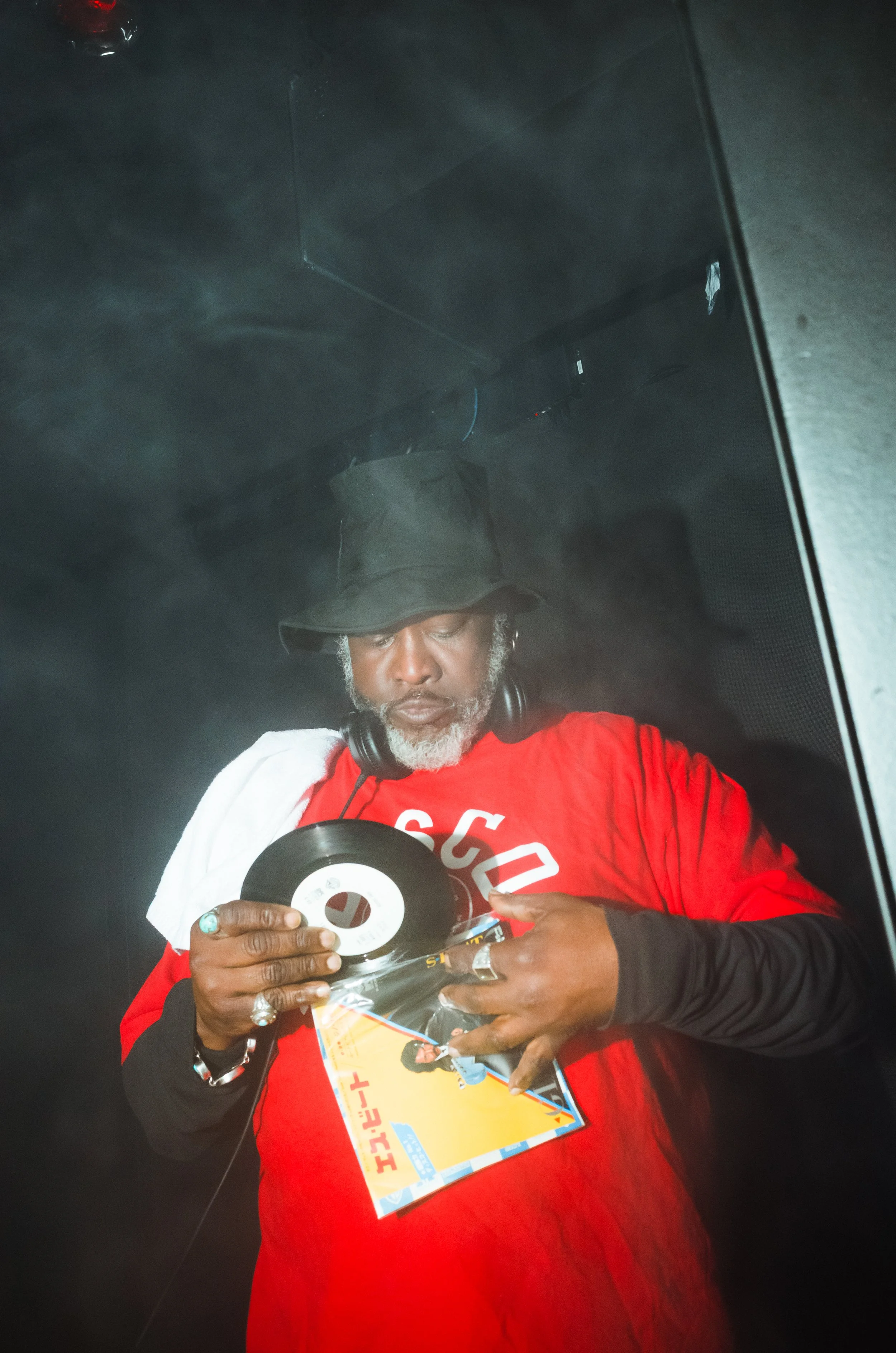 A man in a red shirt, wearing a black hat and headphones, is holding a vinyl record and a magazine with a person on the cover.