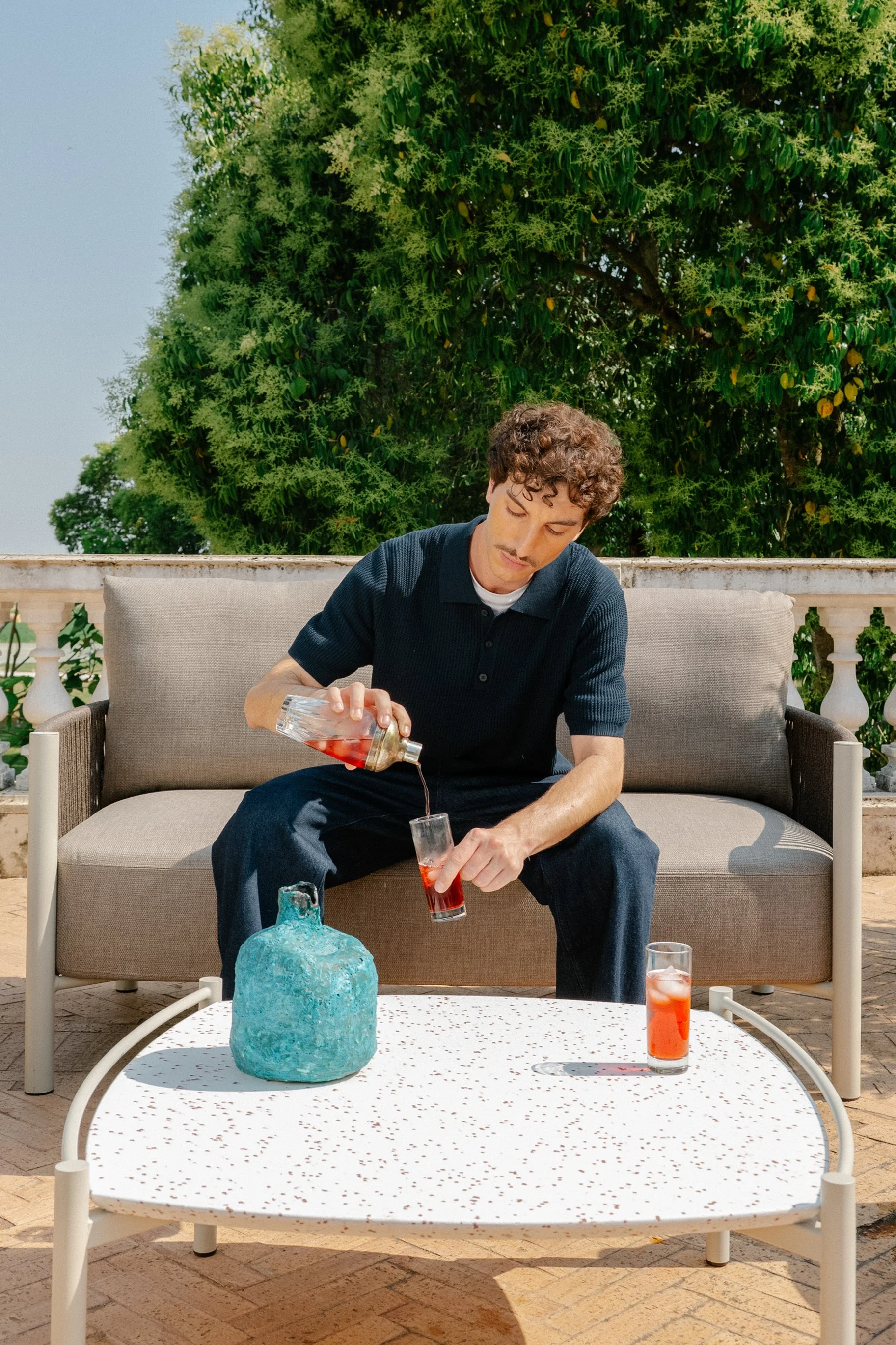 A young man with curly hair wearing a navy blue polo shirt pours a pink cocktail into a glass on an outdoor patio, sitting on a beige sofa with trees in the background.
