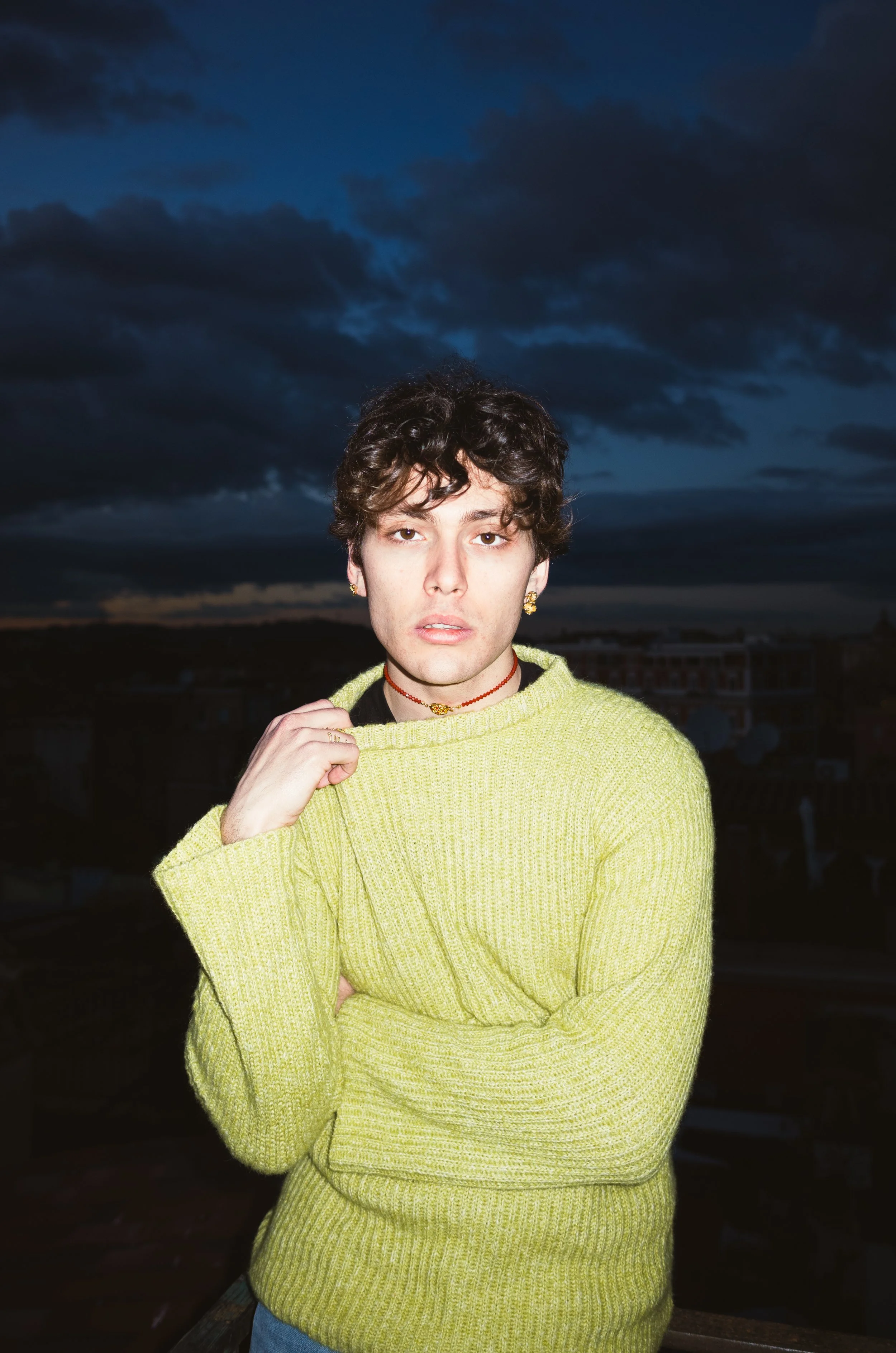A young person with dark curly hair wearing a yellow-green sweater and jewelry, standing outdoors at dusk with dark clouds in the sky.