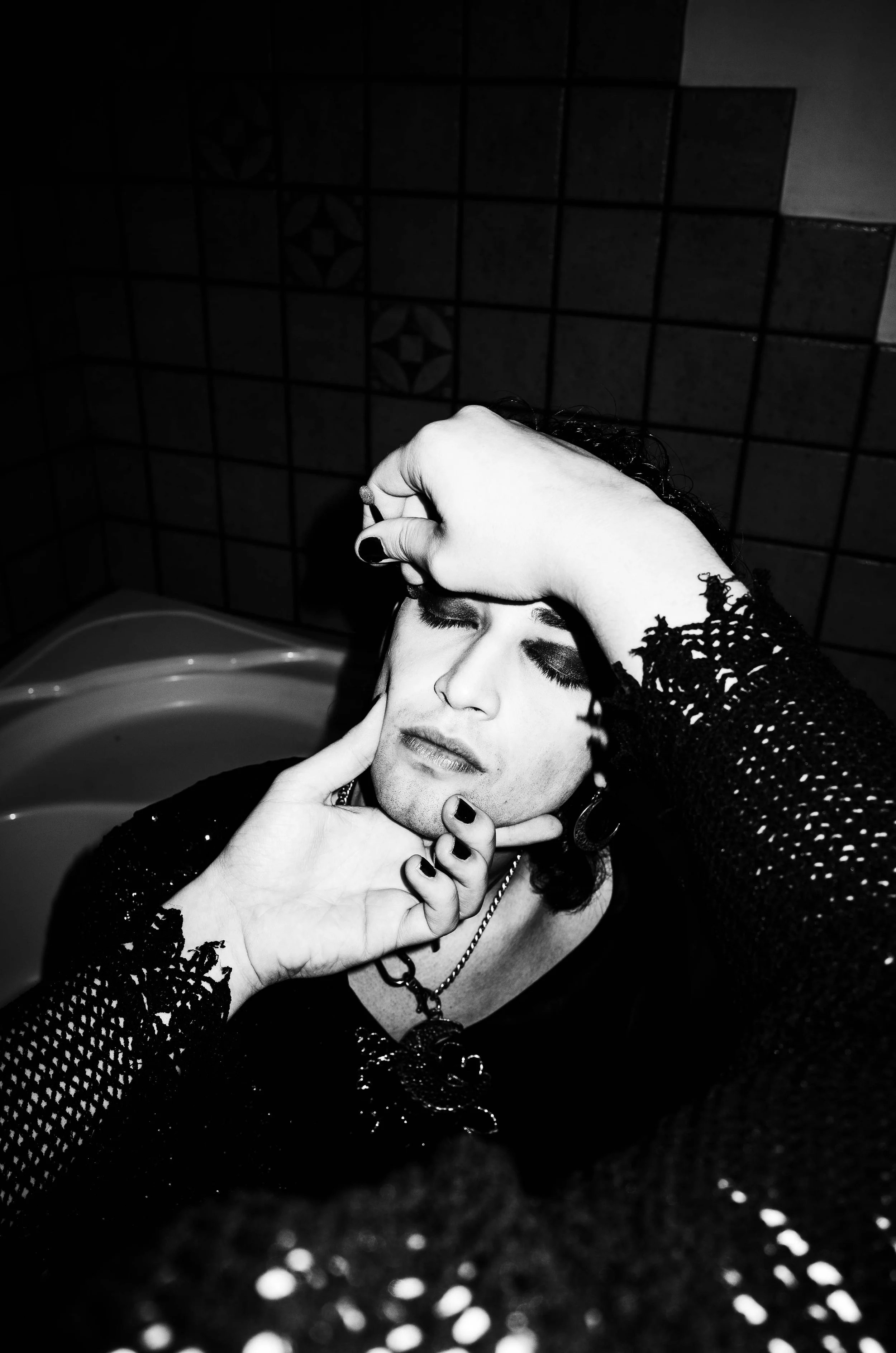 A person with dark eye makeup and jewelry has eyes closed and is being gently held by two hands, one on their forehead and the other on their chin, in a black-and-white photo with tiled wall background.