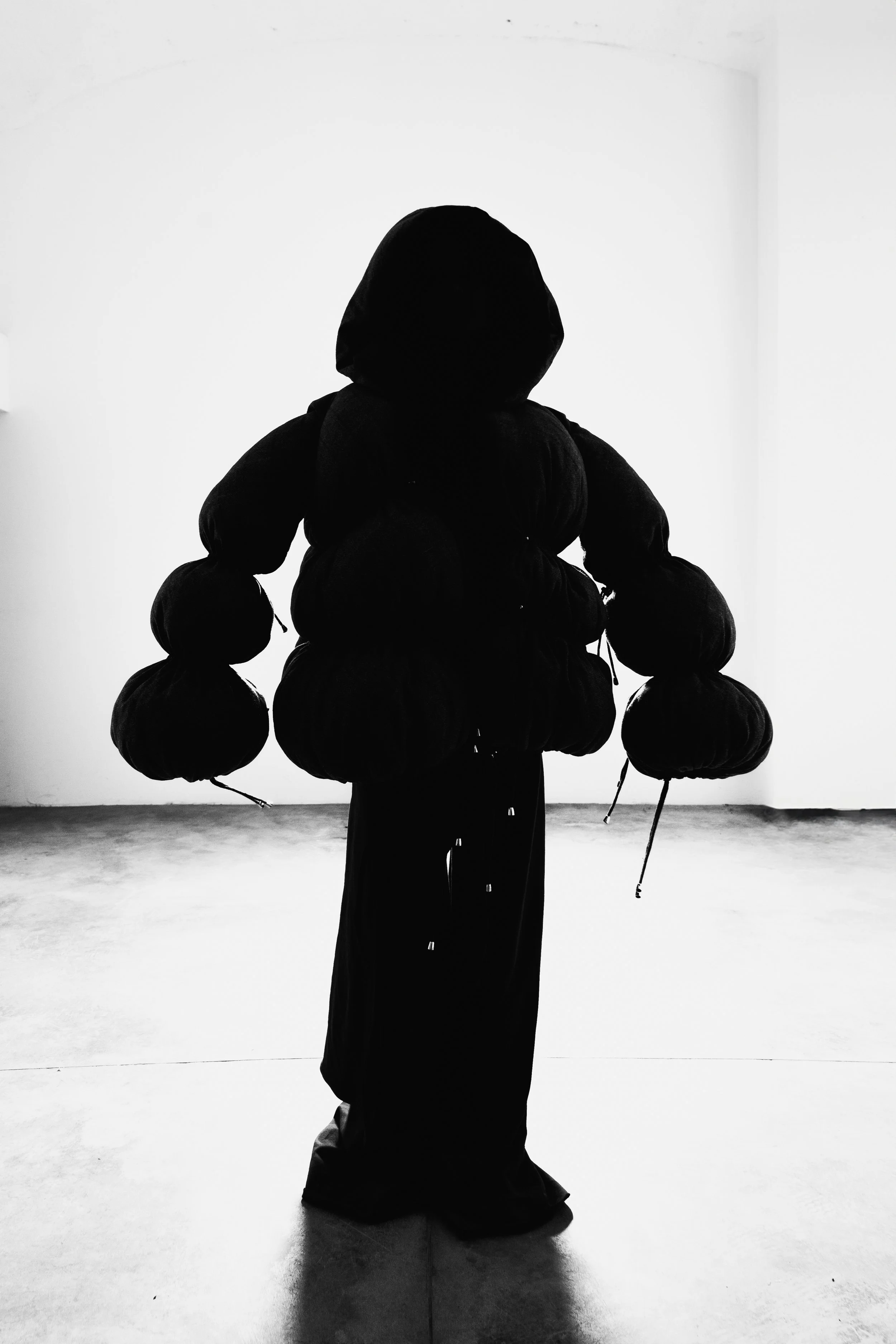 Person dressed in dark clothing and a hood, carrying multiple large black bags, standing against a plain white wall.