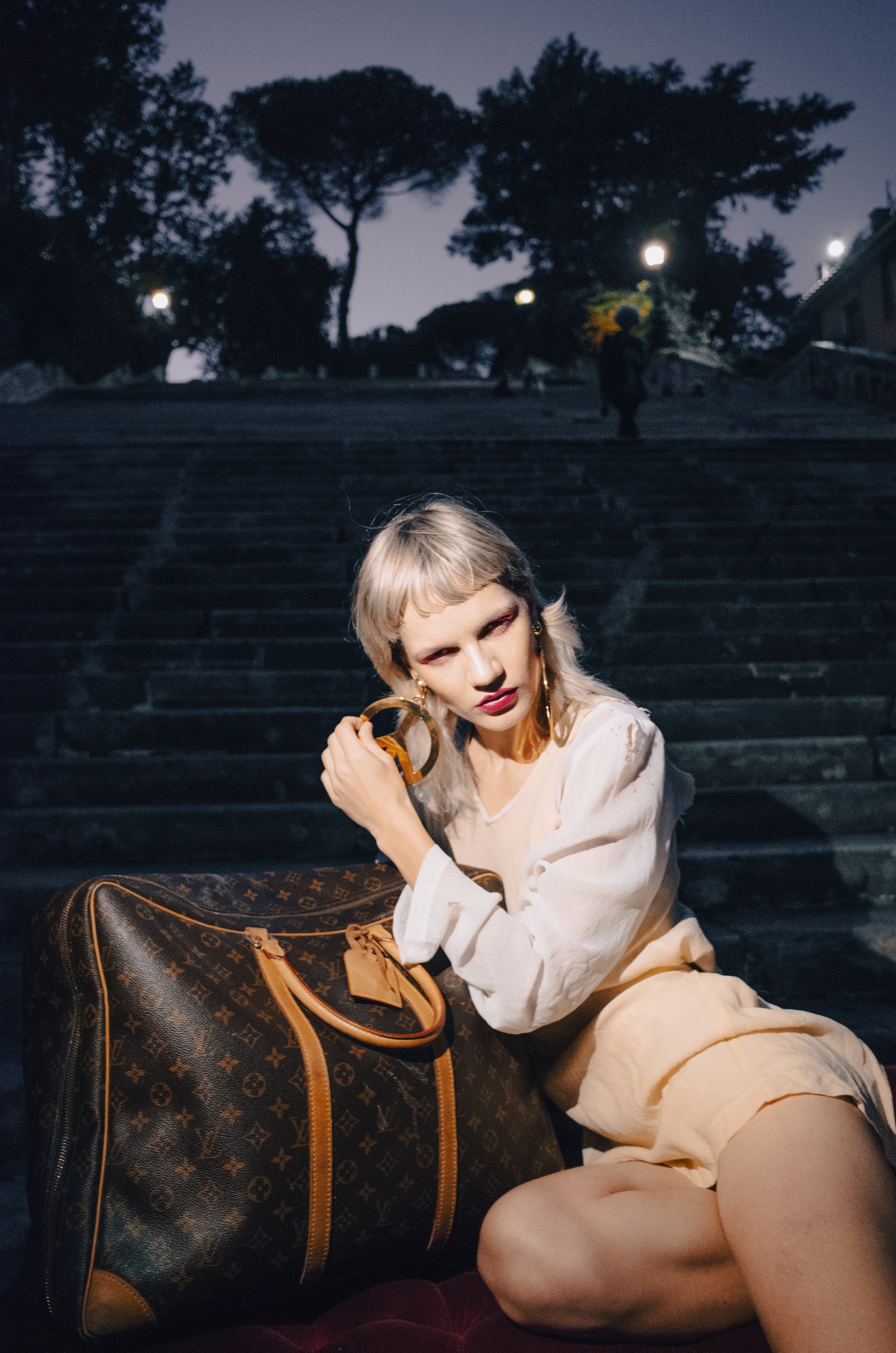 A woman with platinum blonde hair and makeup sitting on stairs at night, holding a large Louis Vuitton bag and wearing large hoop earrings.