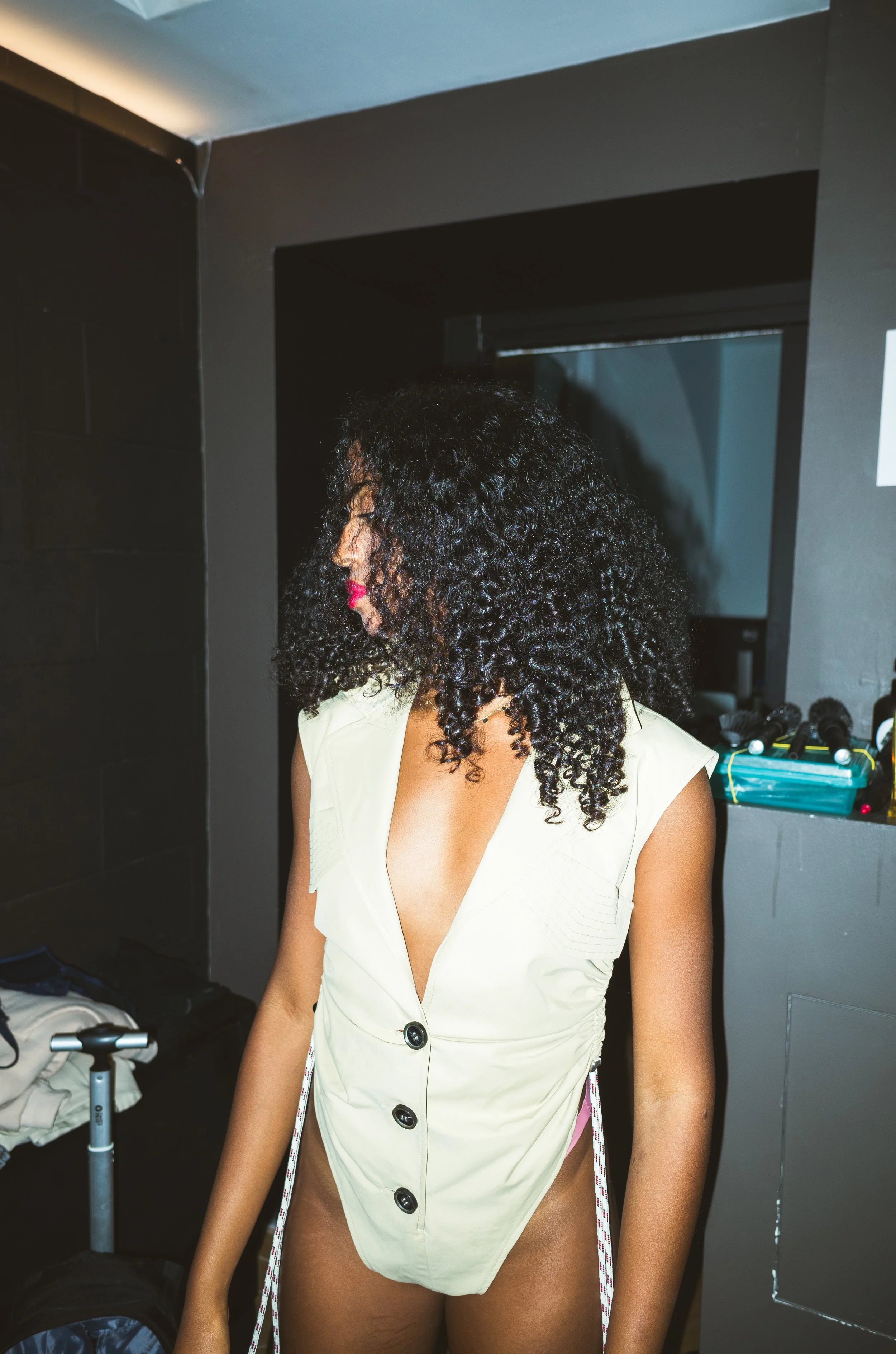 A woman with curly black hair and pink lipstick wearing a cream-colored sleeveless jacket with large buttons, standing in a room with dark walls, near a dressing table with styling tools and hairbrushes, looking downward.