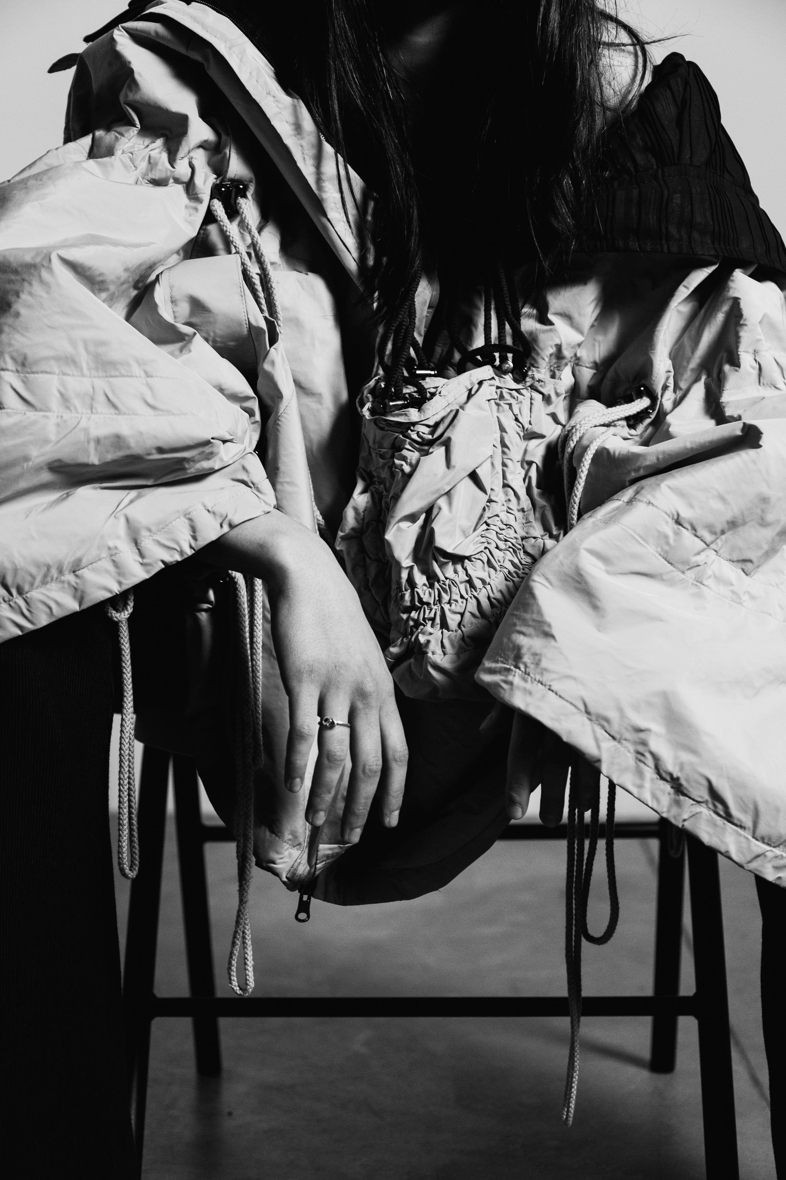 A black and white photo of a person wearing a large, puffy jacket. The person's face is not visible, and they are seated with their hand resting on their lap, showing a ring on their finger.