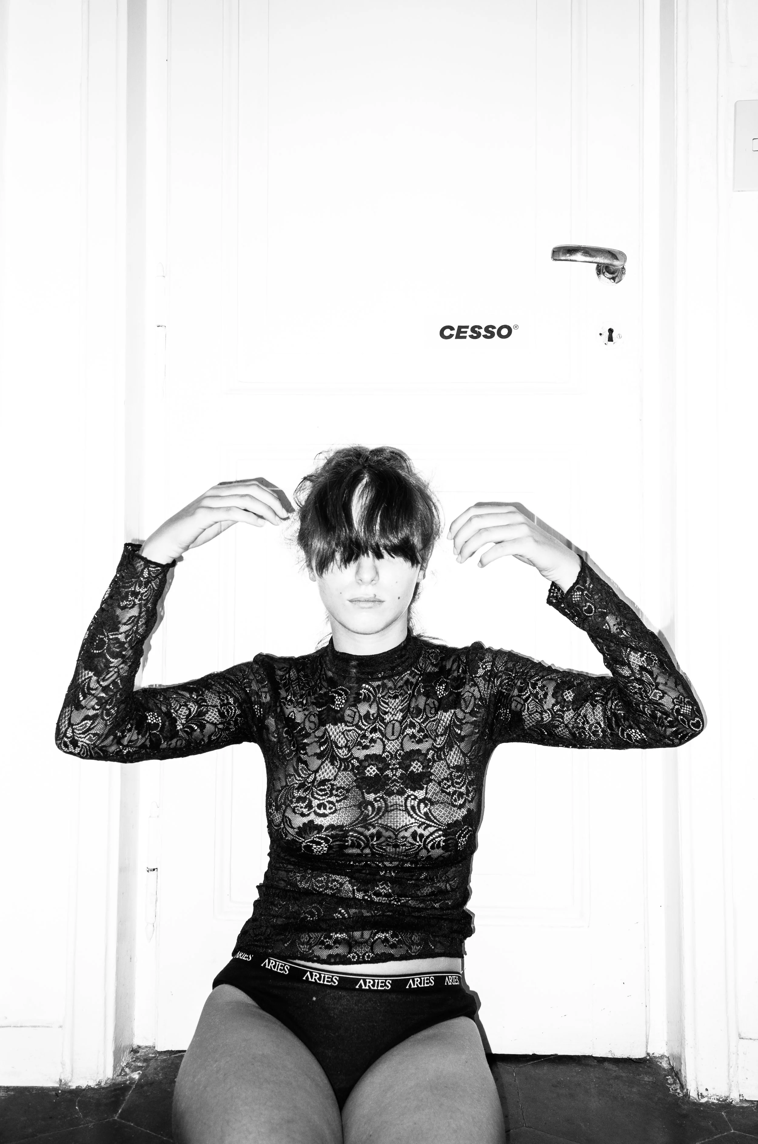 A woman with short dark hair covering her eyes, wearing a black lace top and black can-Can underwear with 'Aries' written on the waistband, sitting on the floor against a white door with the brand 'Cesso' near the handle.