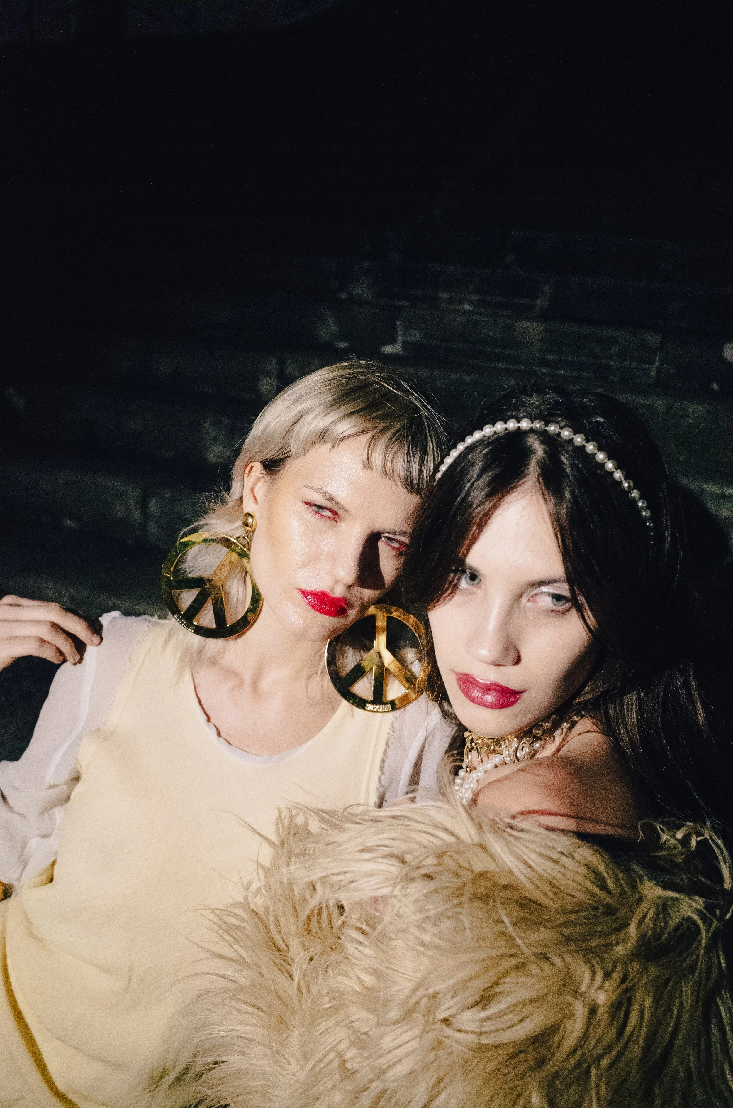 Three women with bold makeup and accessories, including peace sign earrings and a pearl headband, taking a close-up selfie.