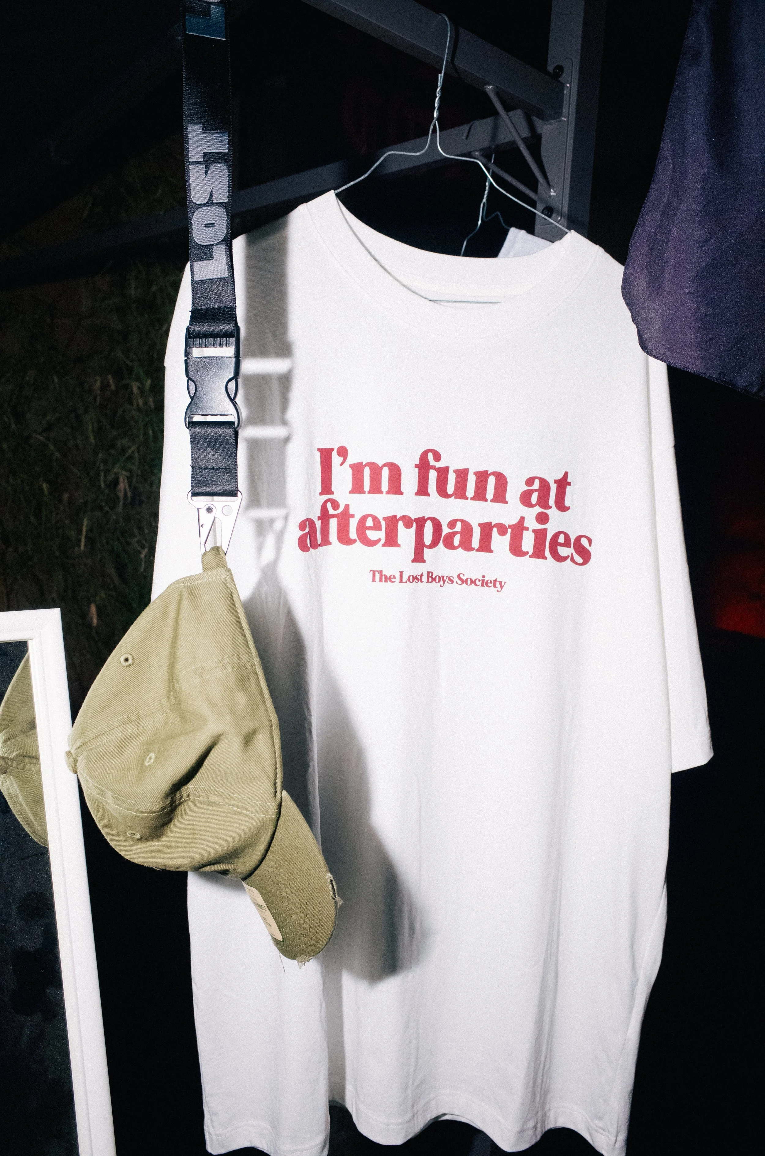 White T-shirt with red text that reads, "I'm fun at afterparties" and smaller text below that says, "The Lost Boys Society." A beige cap and a black strap with the word "LOST" are hanging on a rack.