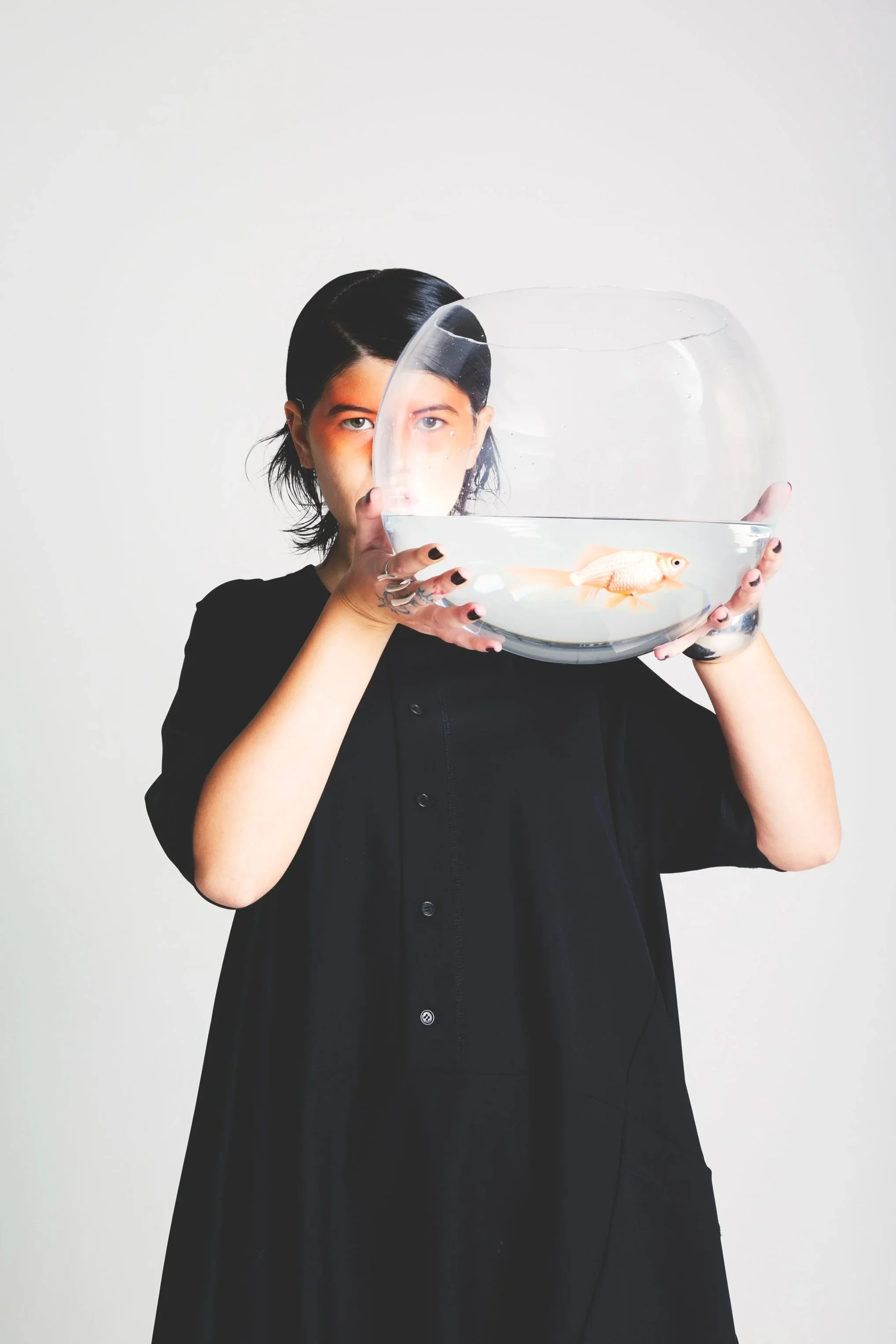 A person with dark hair holding a large fishbowl with a goldfish inside, partially obscuring their face.