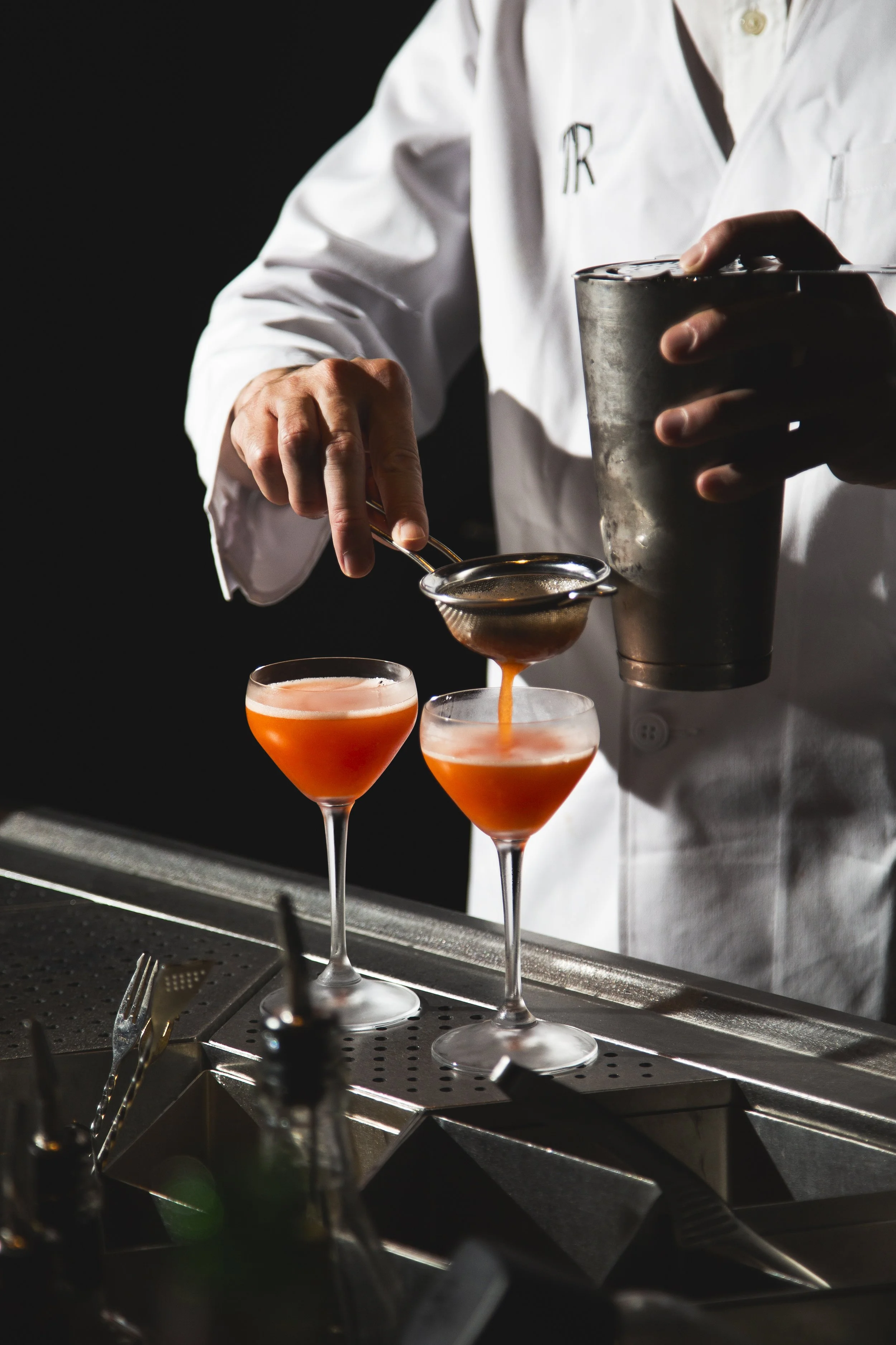 A bartender in a white shirt pours a cocktail through a strainer into two coupe glasses with pinkish-orange drinks. The background is dark, and the bartender holds a shaker in one hand.