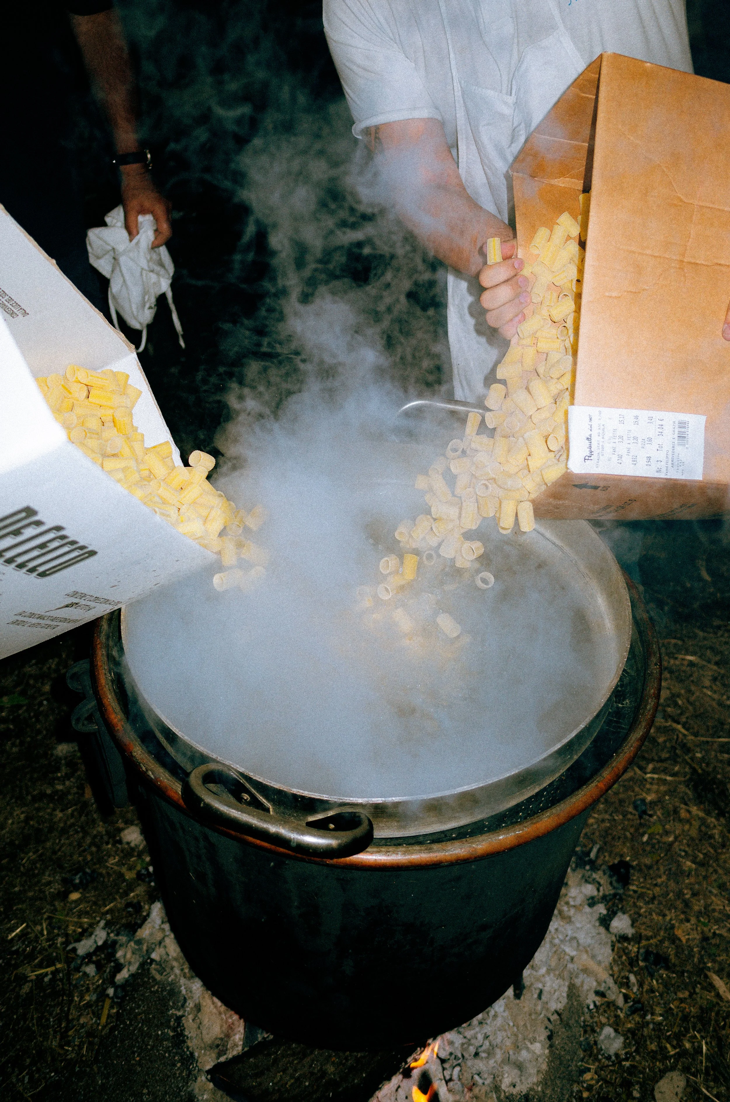 Two people pour large quantities of yellow pasta from boxes into a steaming pot over open flame outdoors.