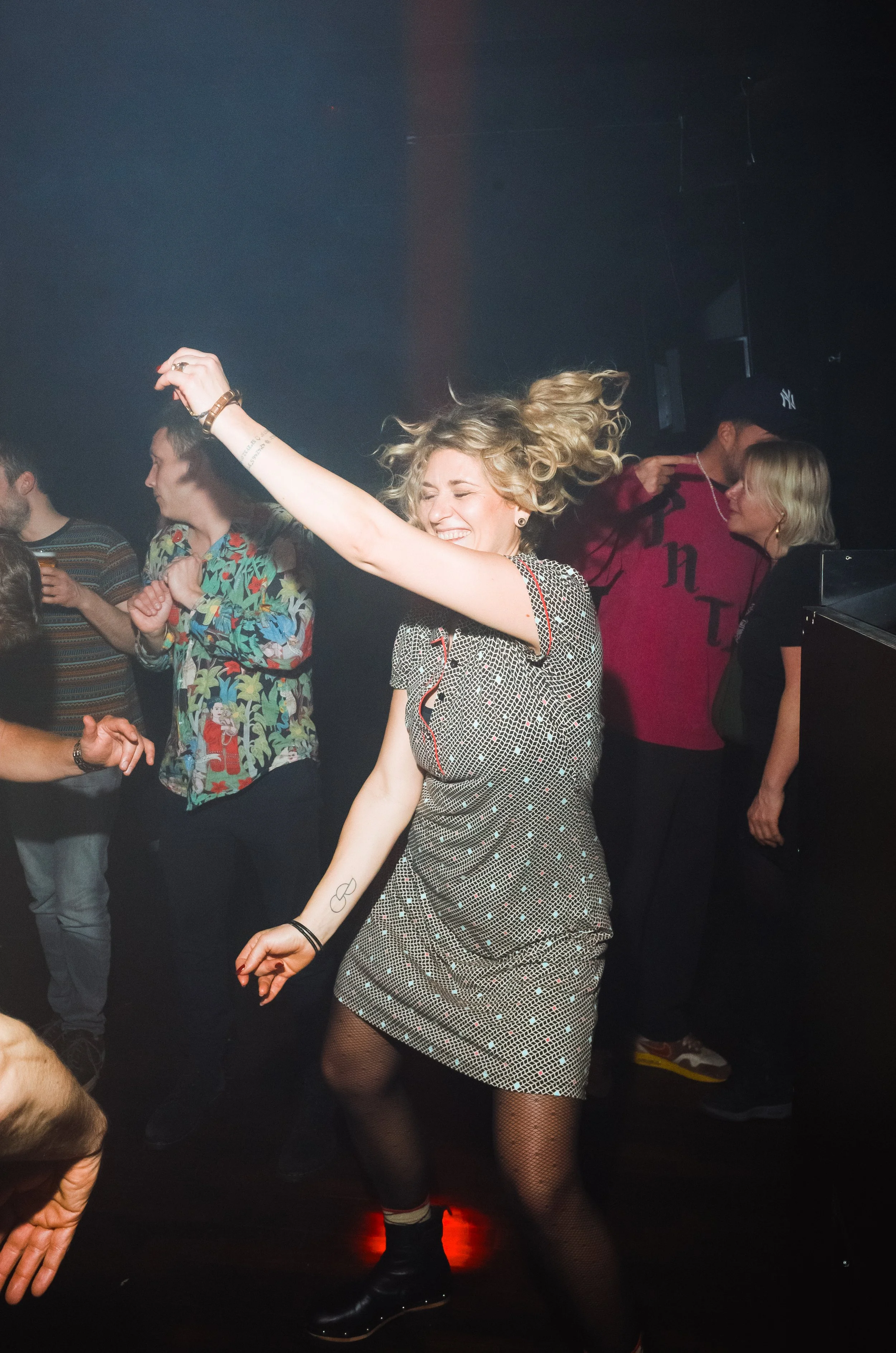 A woman dancing happily in a nightclub with her arm raised, surrounded by other people. She is smiling with her eyes closed, wearing a silver patterned dress, fishnet stockings, and ankle boots.