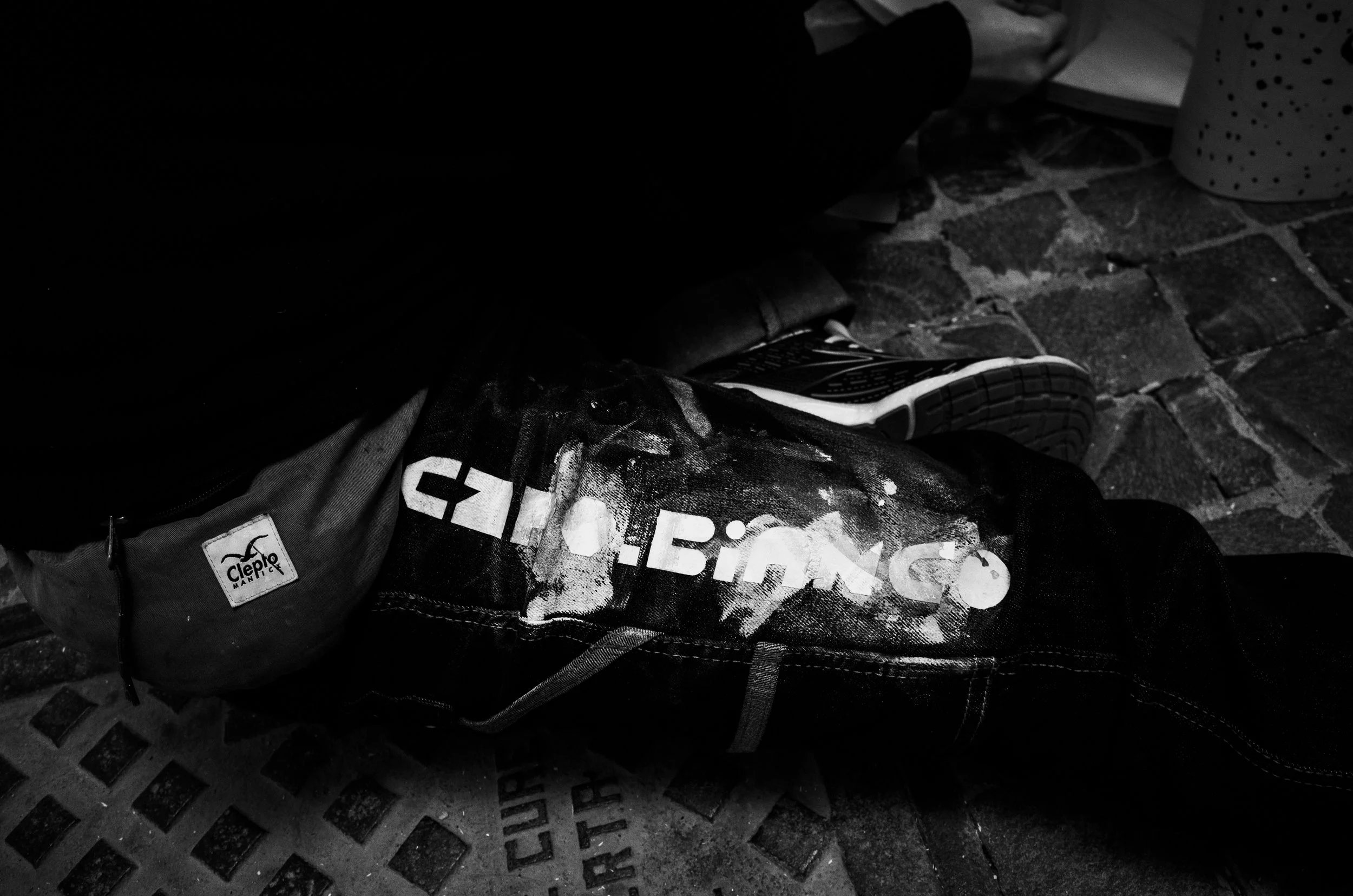 Black and white photo of a discarded backpack and a pair of sneakers on a brick sidewalk.