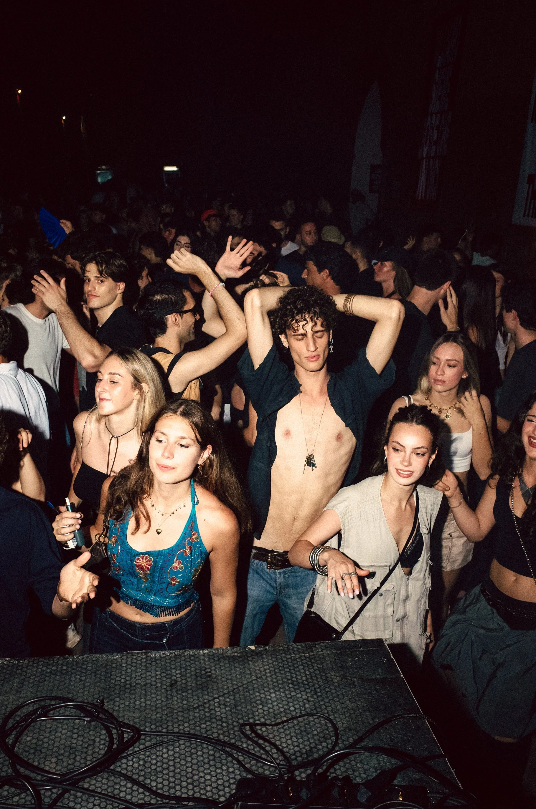 Crowd of young people dancing at a concert or party, with a person in the center with curly hair, shirt open, and eyes closed, surrounded by others enjoying the music.