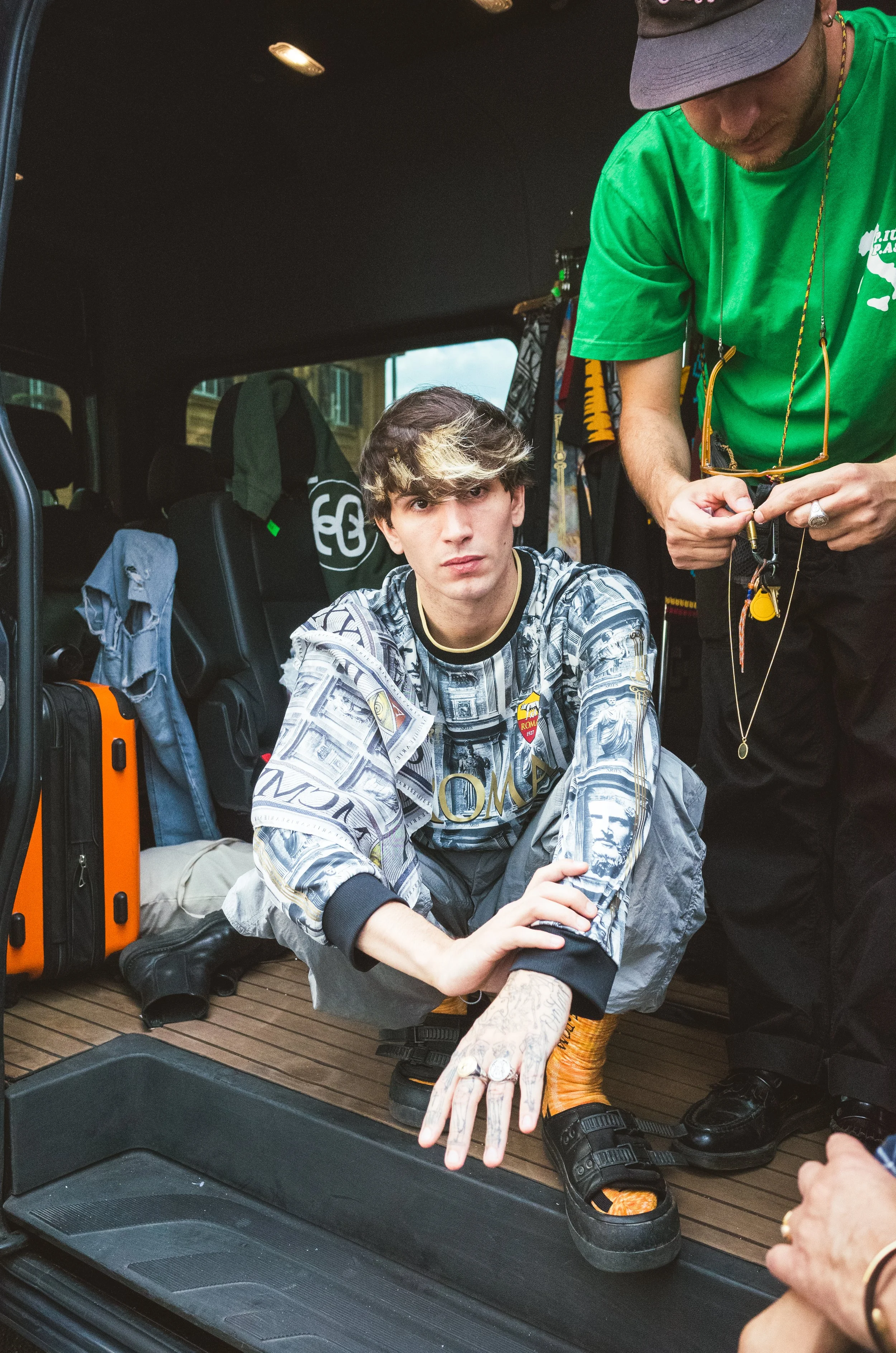 A young man with tattoos and styled hair sitting inside a vehicle, looking at the camera, with luggage and backpacks around him.