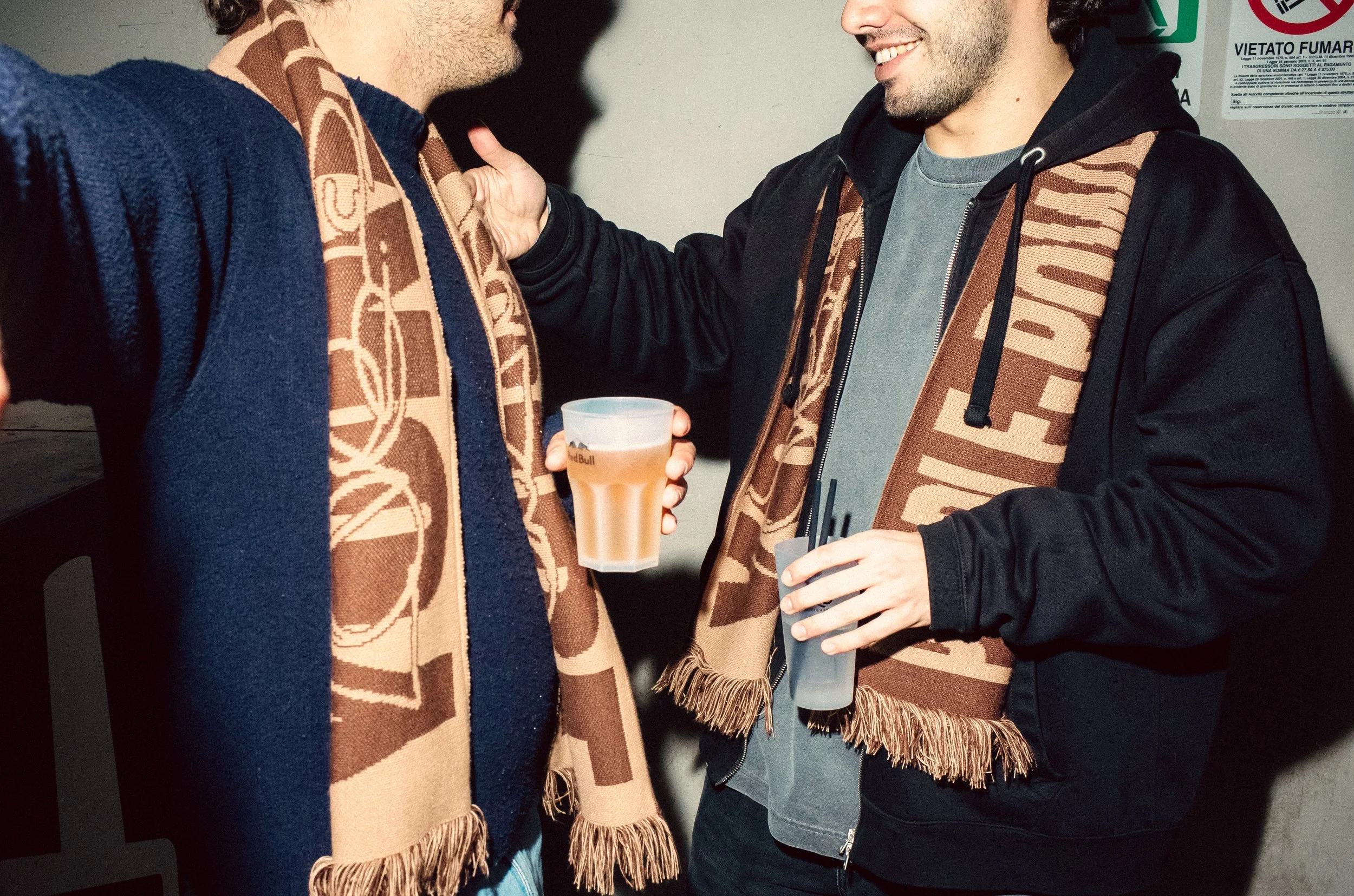 Two men standing close together, wearing brown scarves with a logo, holding drinks, and smiling at each other.