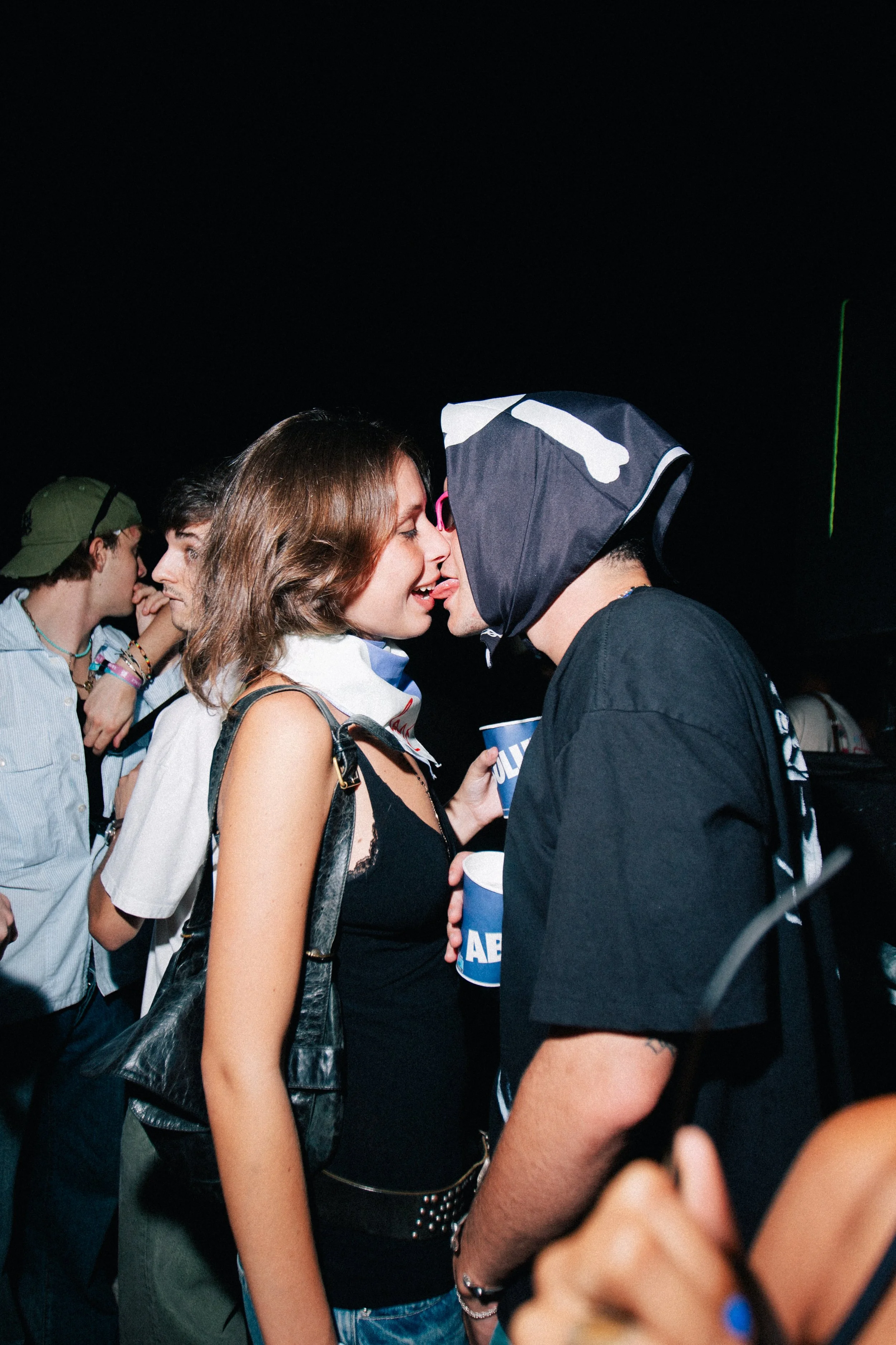Two young people, a woman and a man, are close together, appearing to be about to kiss, with other people in the background at a crowded event or party.