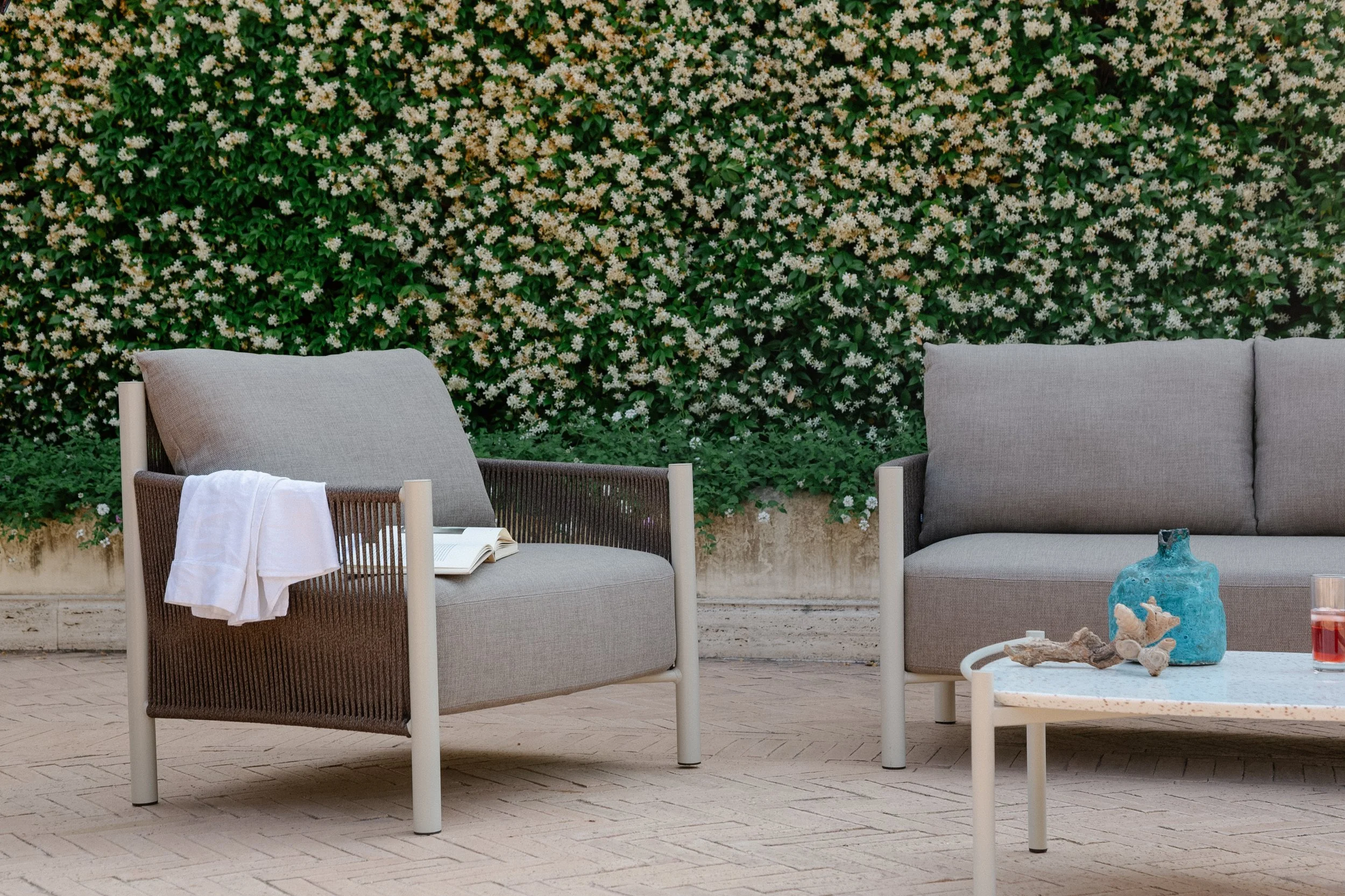 Outdoor seating area with two modern gray armchairs and a gray sofa, a white coffee table with decorative objects including a blue vase and driftwood, and a lush green hedge with white flowers in the background.