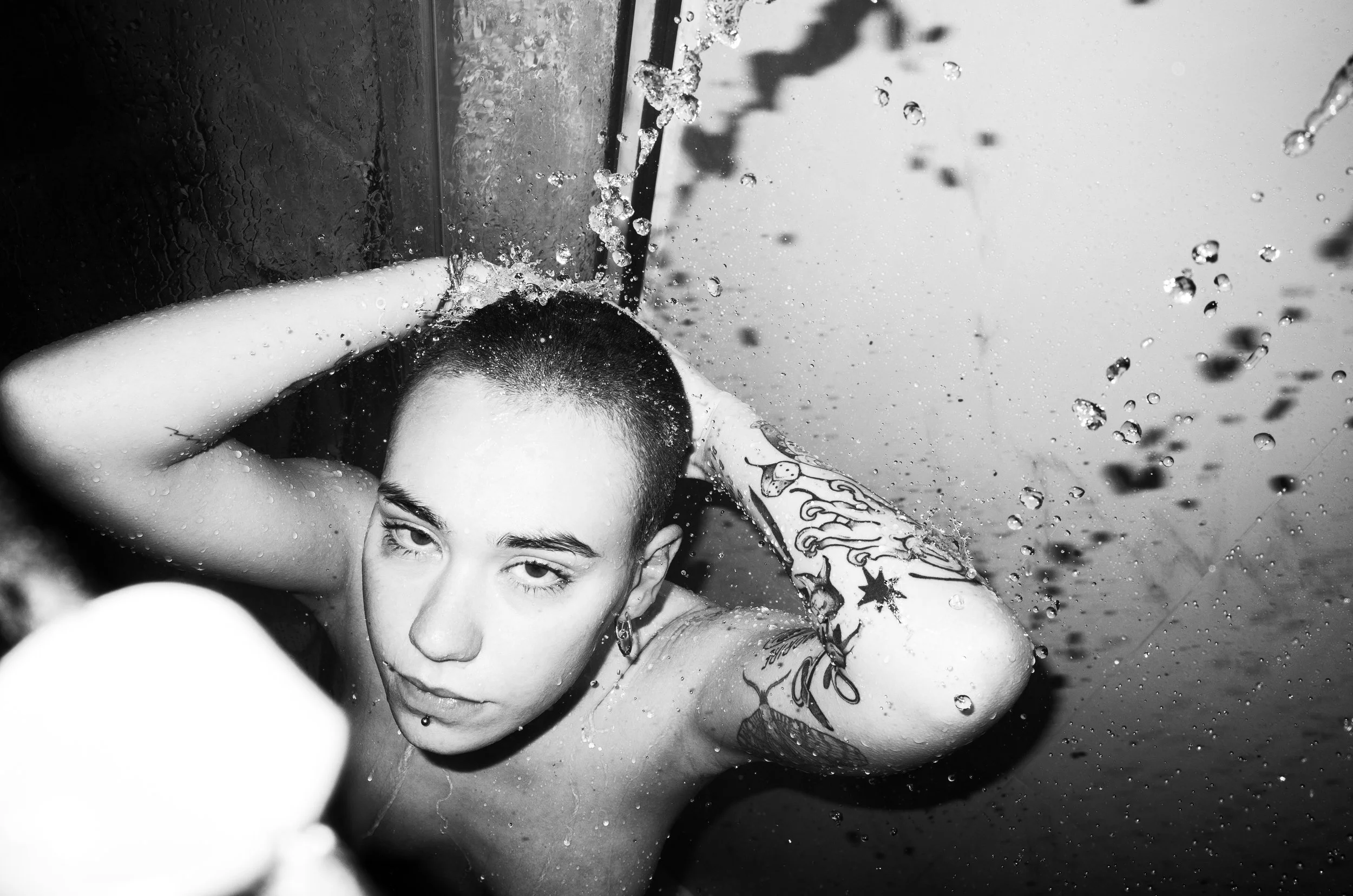 A person with short hair and tattoos on their arms, in a shower, with water splashing, looking at the camera.
