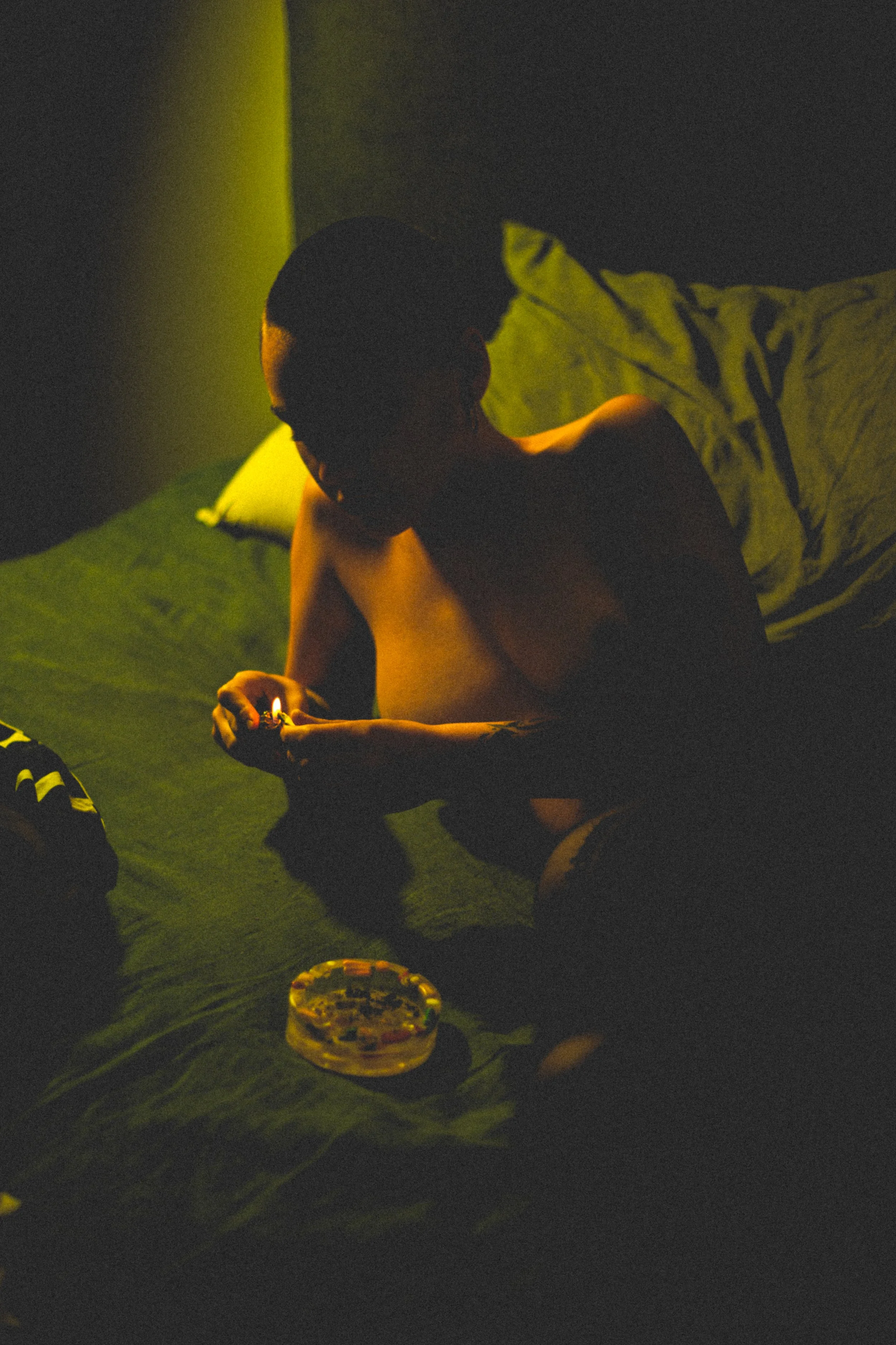 A person with short hair, sitting shirtless on a bed with green sheets, is lighting a cigarette in a dimly lit room.