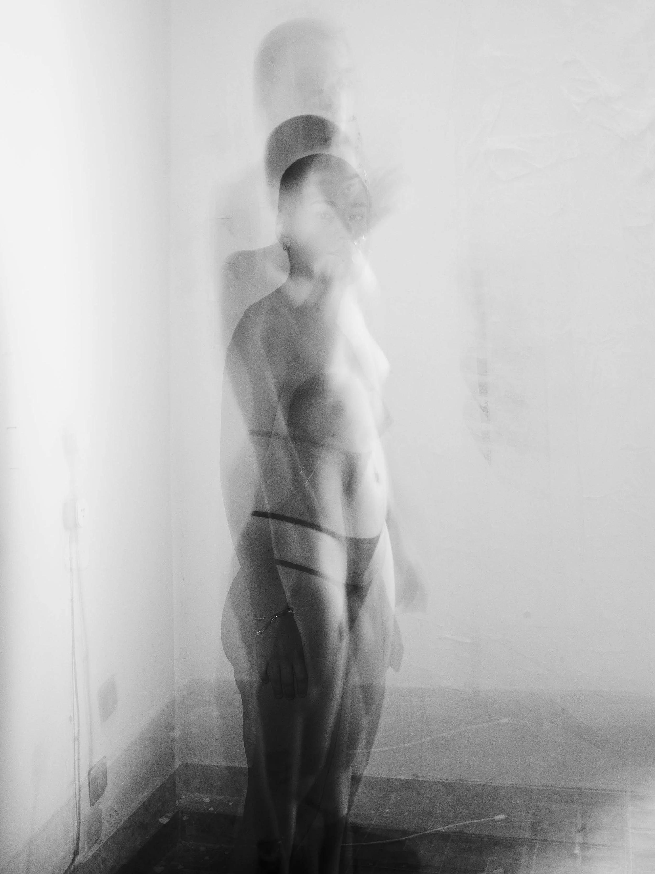 A black and white photograph of a woman standing in front of a plain wall, with multiple faint overlapping images creating a ghostly, double-exposure effect.