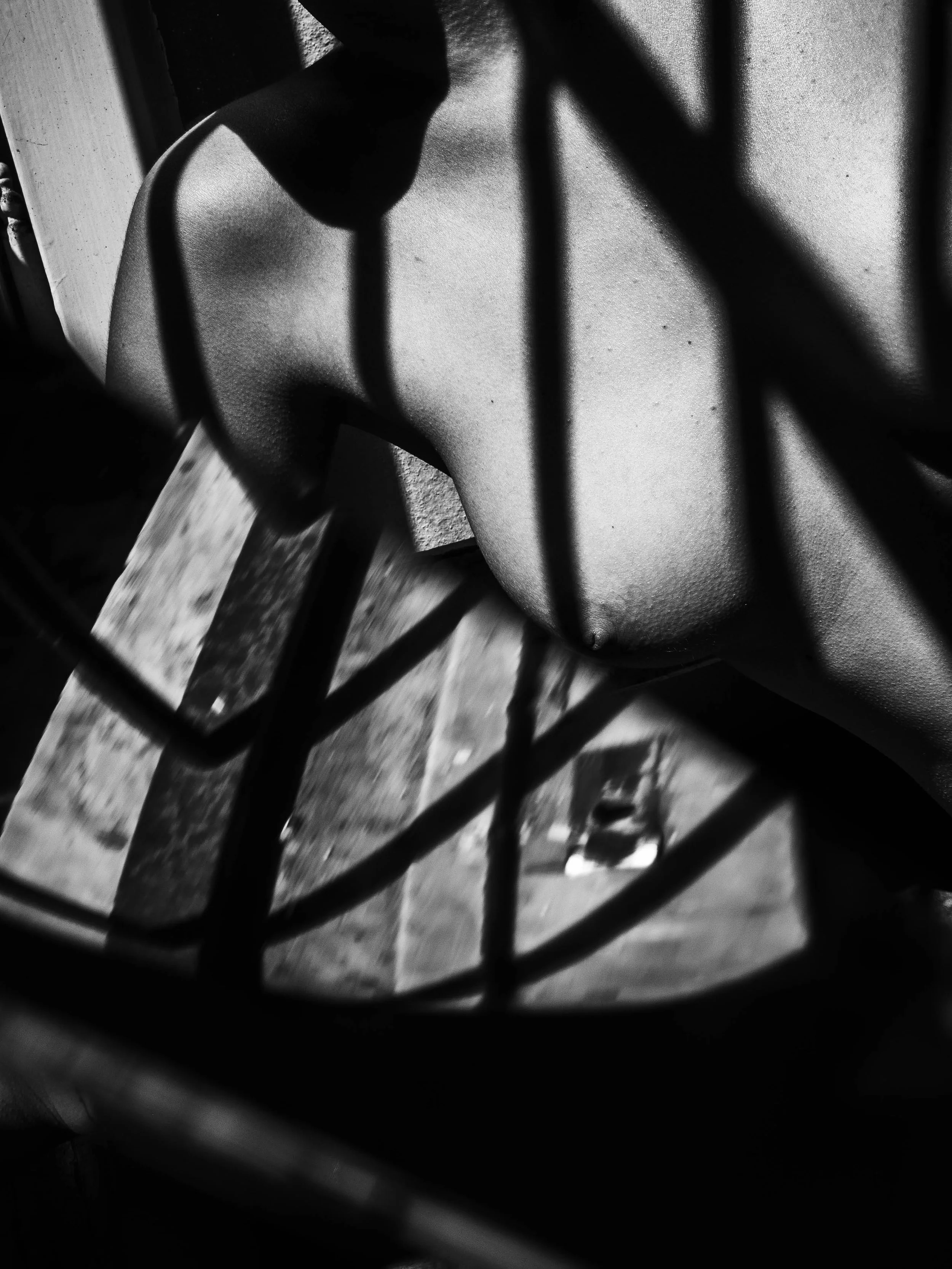 Black and white photo of a person's back and shoulder seen through a metal railing, with sunlight casting shadows.