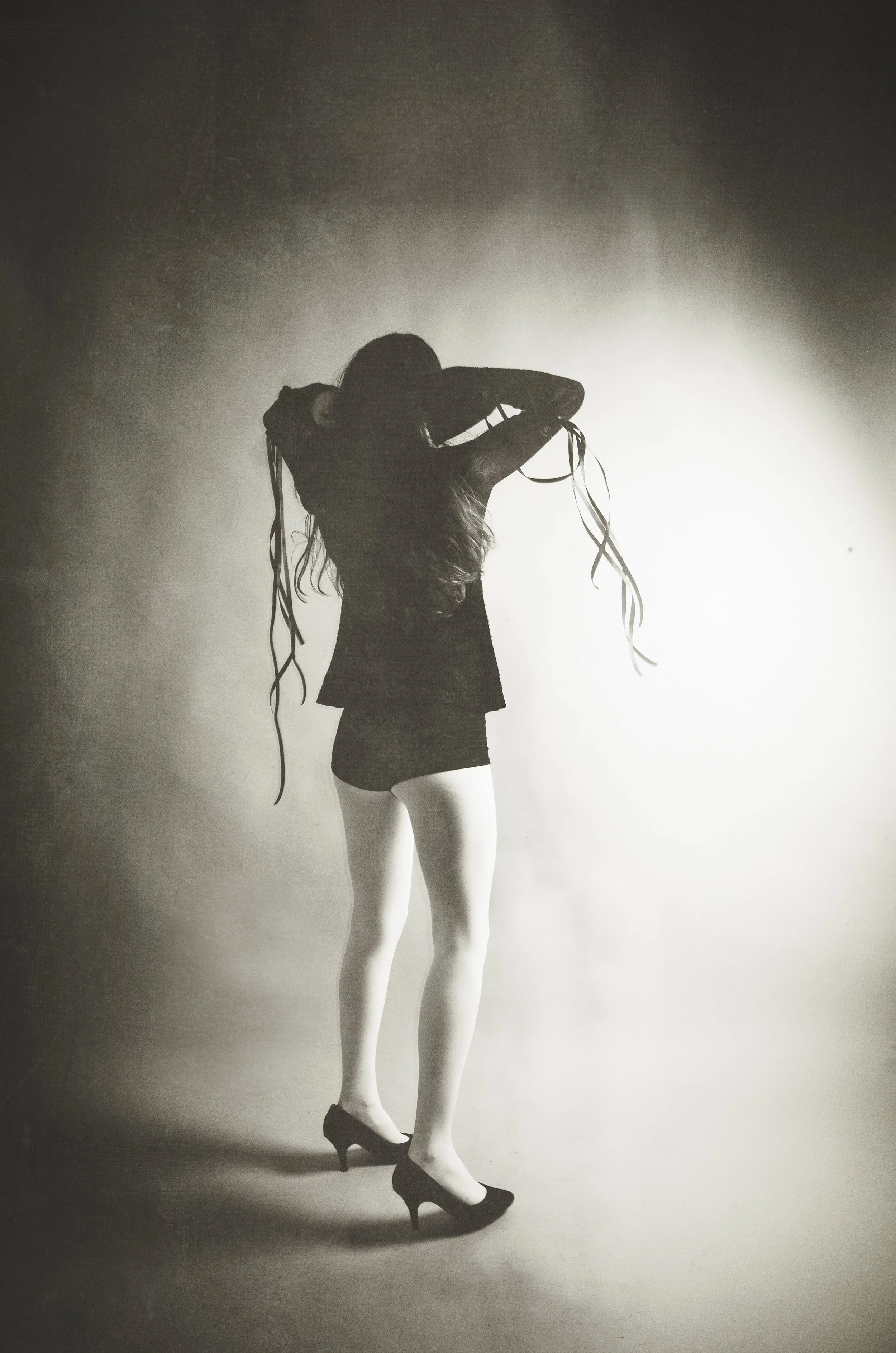 Black and white photo of a woman in high heels, wearing a blazer and shorts, with long hair, standing against a plain background, adjusting her hair.