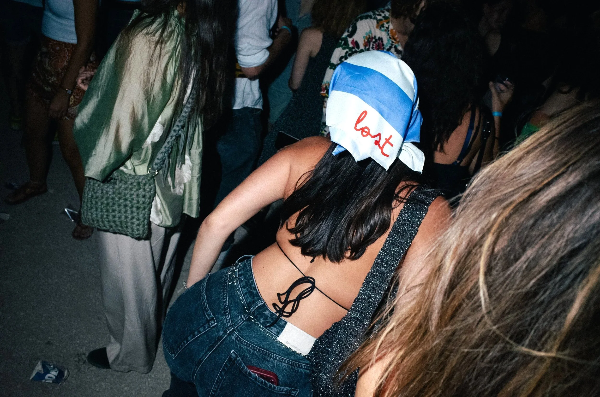 A woman wearing a blue and white bandana with red writing, a black spaghetti strap crop top, and high-waisted blue jeans is in a crowded outdoor setting during the evening or night.