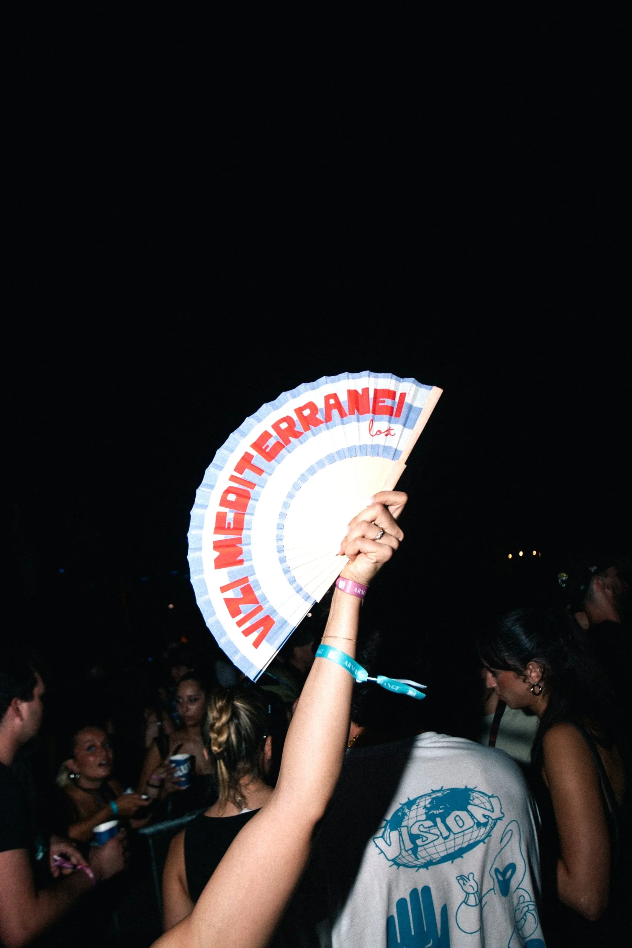 Person holding a fan with red and blue text at a nighttime outdoor event, surrounded by people.