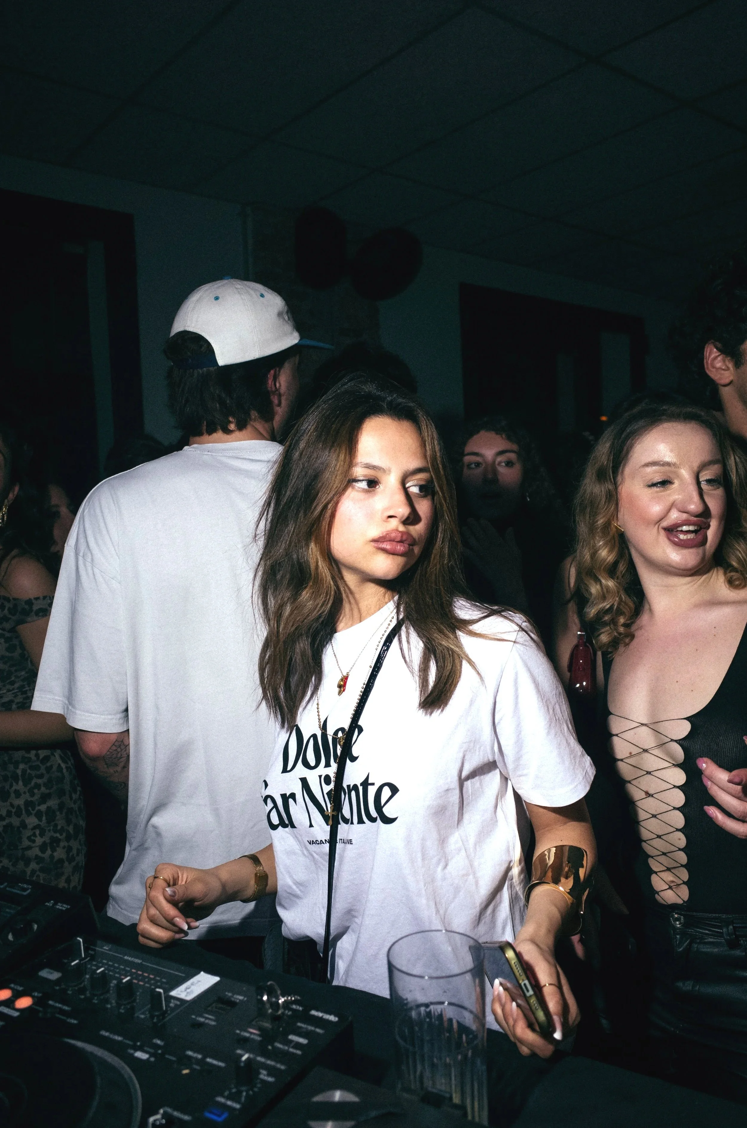 A young woman with long brown hair wearing a white t-shirt, gold jewelry, and holding a smartphone and drink, DJing at a party.