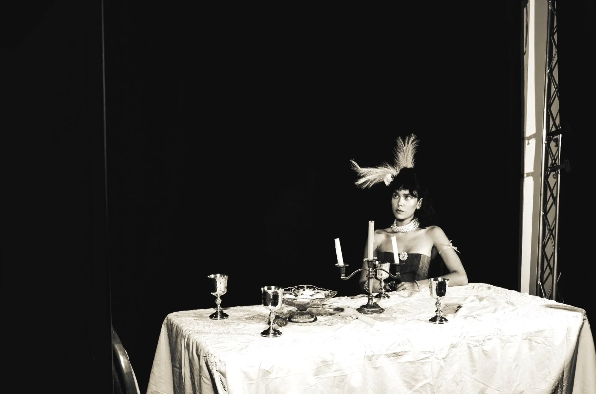 A woman with dark hair wearing a feathered headpiece, pearl necklace, and elegant dress, sitting at a table with a white tablecloth, candelabra, and multiple silver goblets, against a dark background.