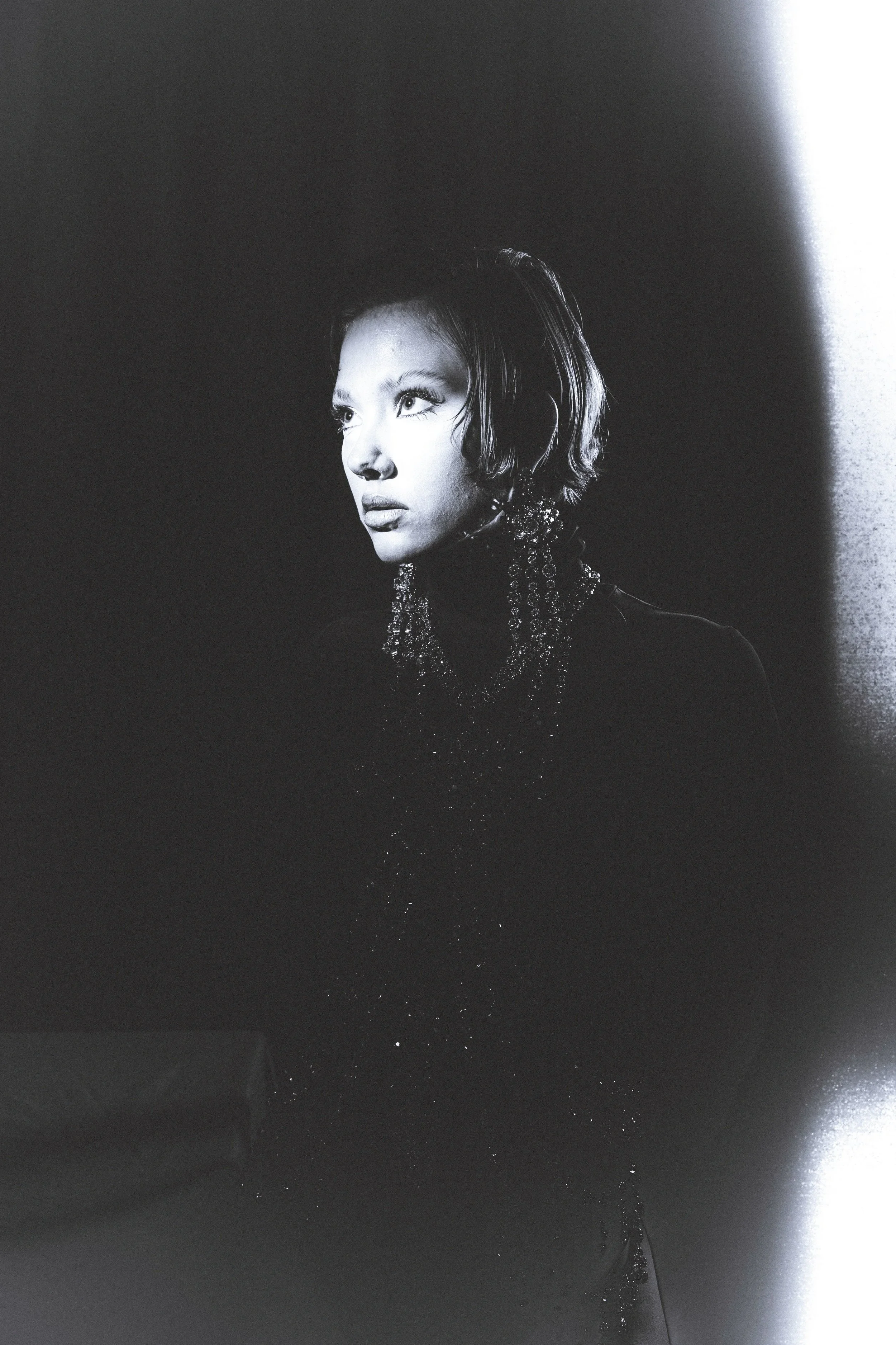 Black and white portrait of a woman with short hair and statement jewelry, looking to the side, with a light source illuminating her face and a dark background.