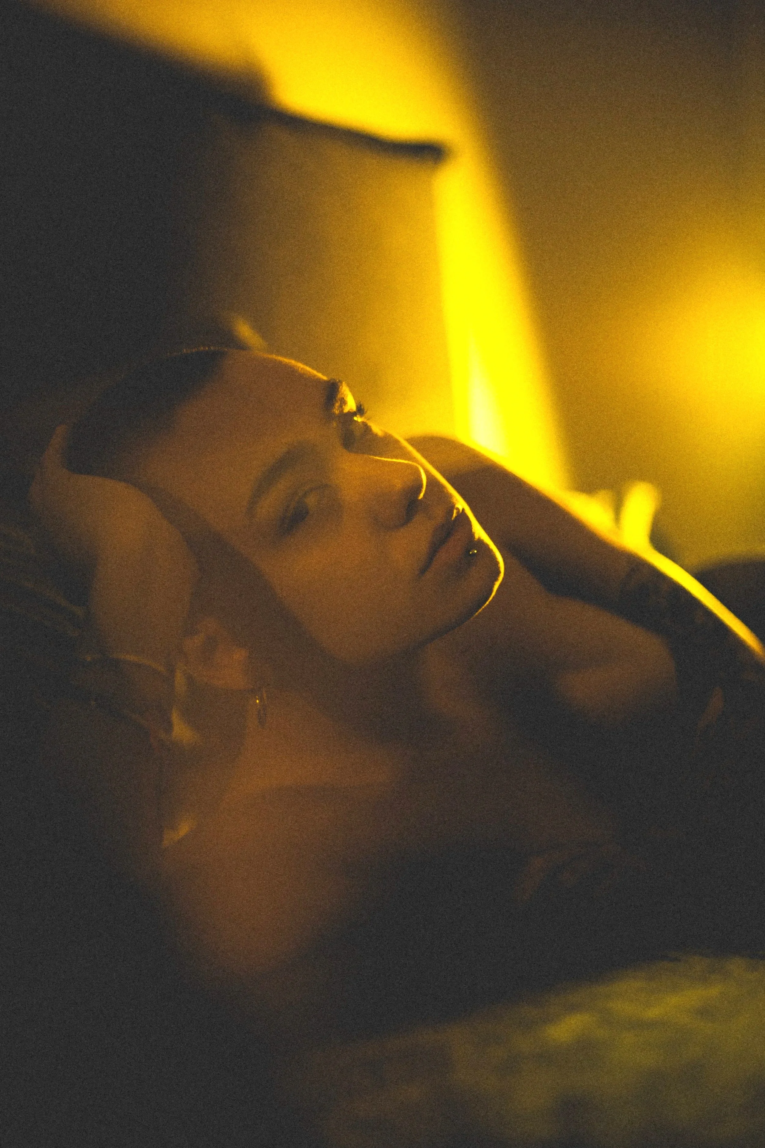 A young woman with short hair lying down in a dimly lit room, illuminated by warm yellow light, with her hand resting on her head.