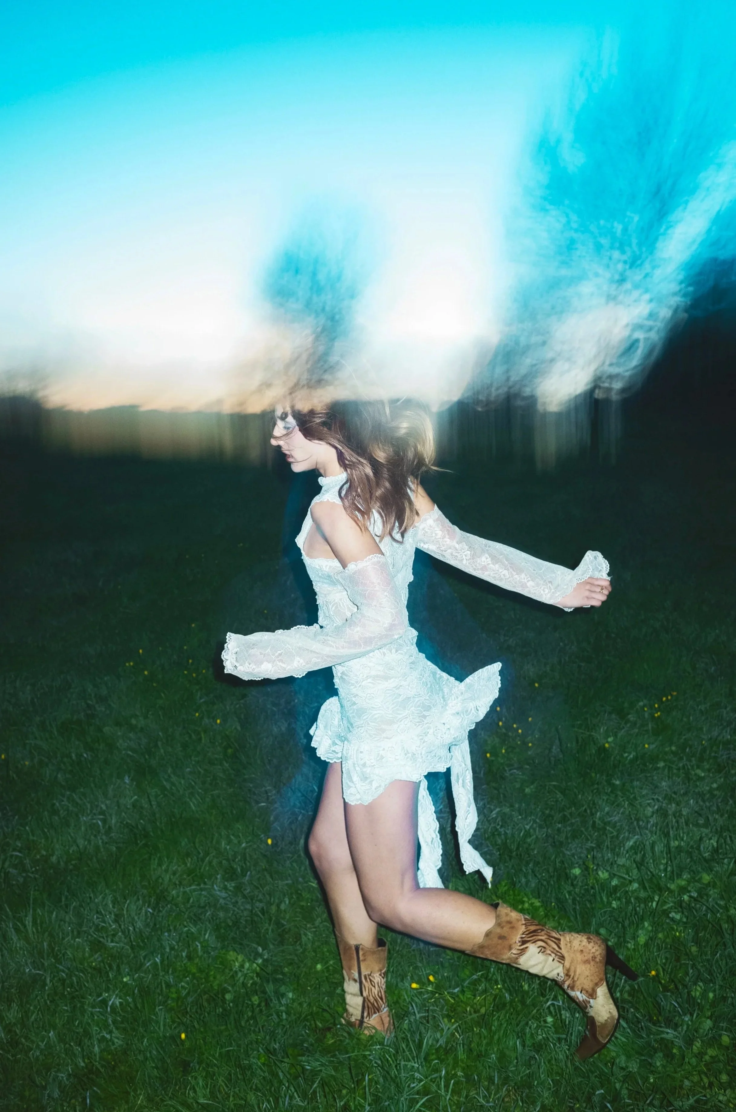 A woman in a white lace dress and brown animal print boots running on a grassy field at dusk or dawn, with a light streak or blurred effect above her head.