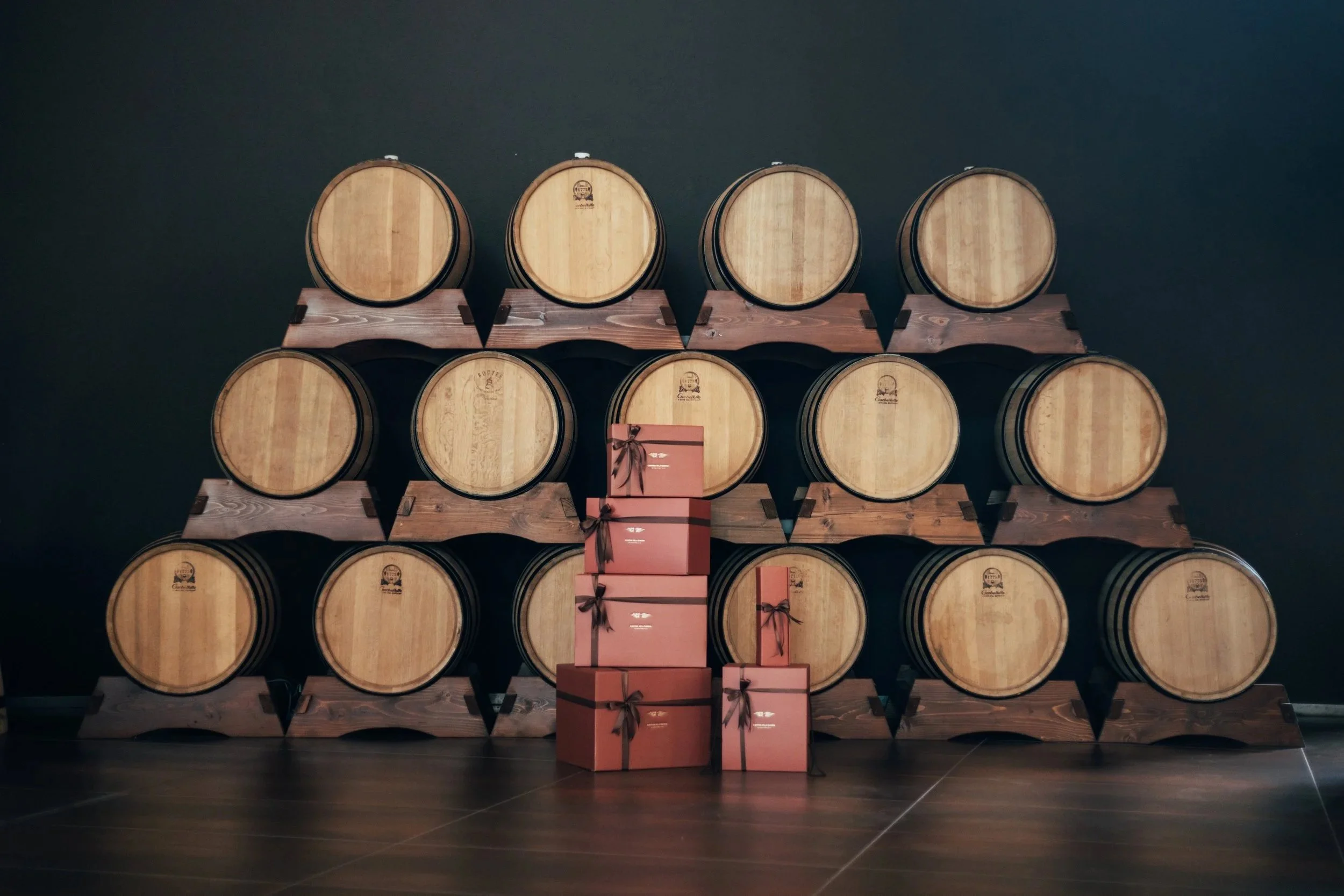 A display of wooden wine barrels stacked on wooden stands with pink gift boxes in front. The background is a dark wall.