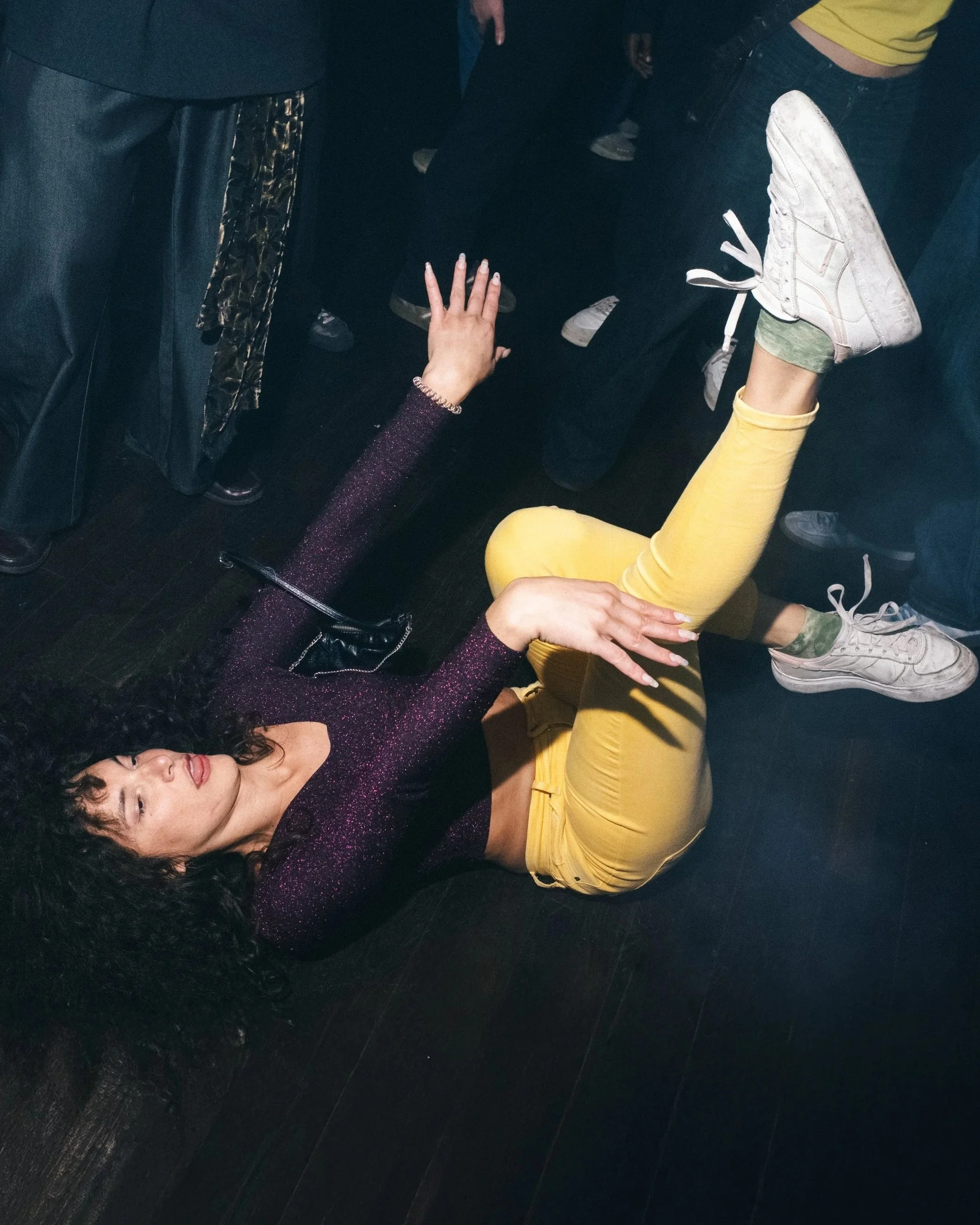 A woman with curly black hair lies on the dark floor, reaching upward with her left hand, surrounded by people at a social event.