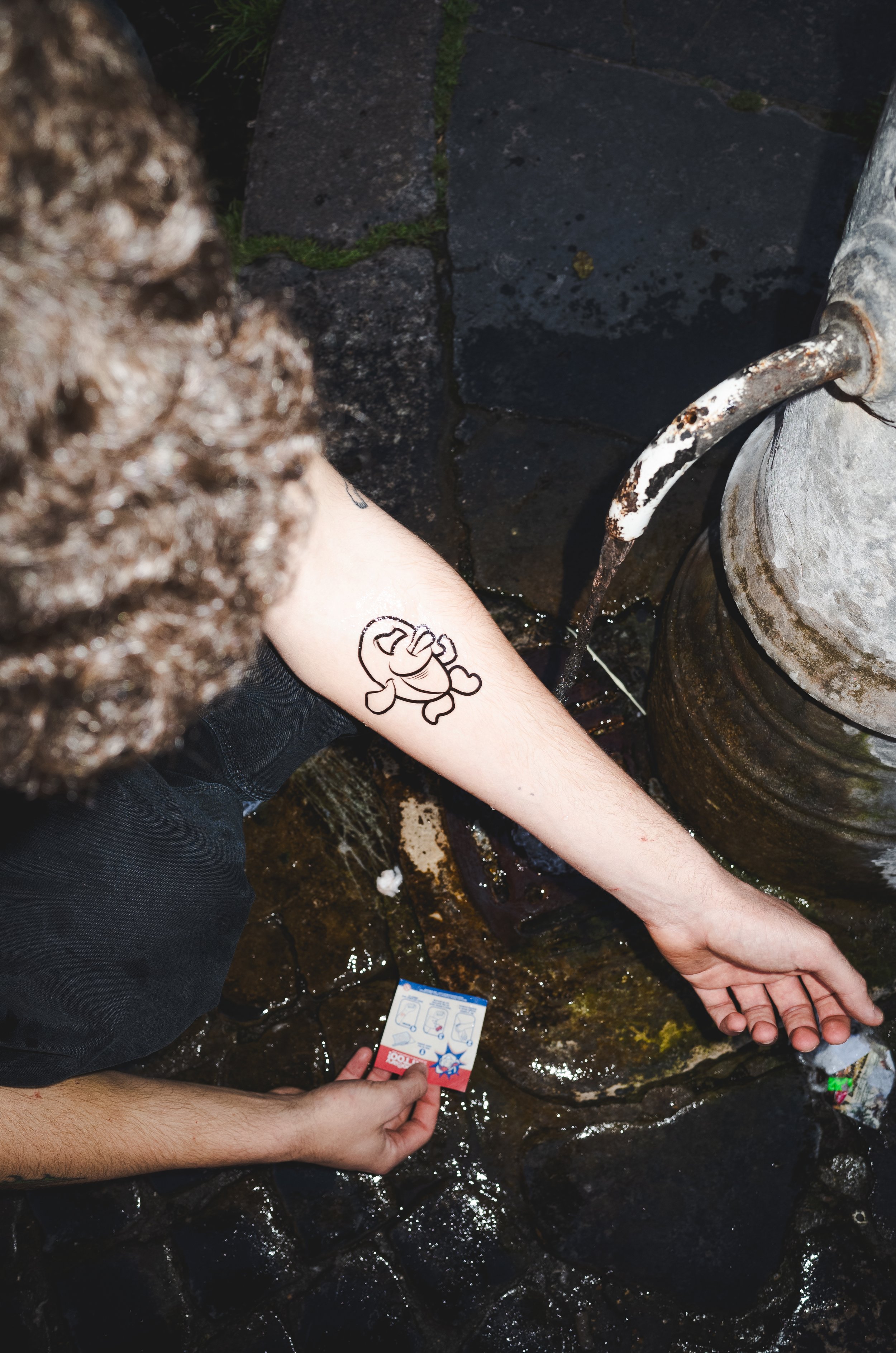 Person with a cartoon tattoo of a character on their arm, filling a plastic bottle with water from a faucet outdoors.