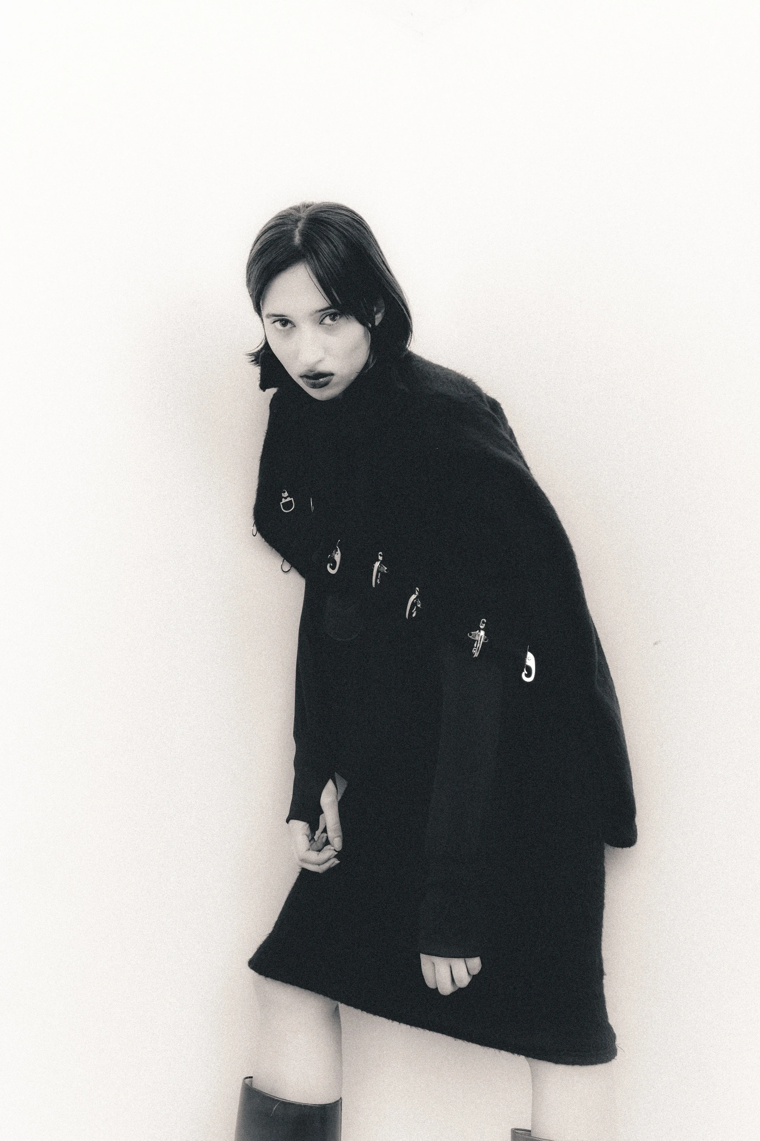 Black and white photo of a woman with dark hair, wearing a dark coat with metal rings, standing against a plain wall and looking at the camera.