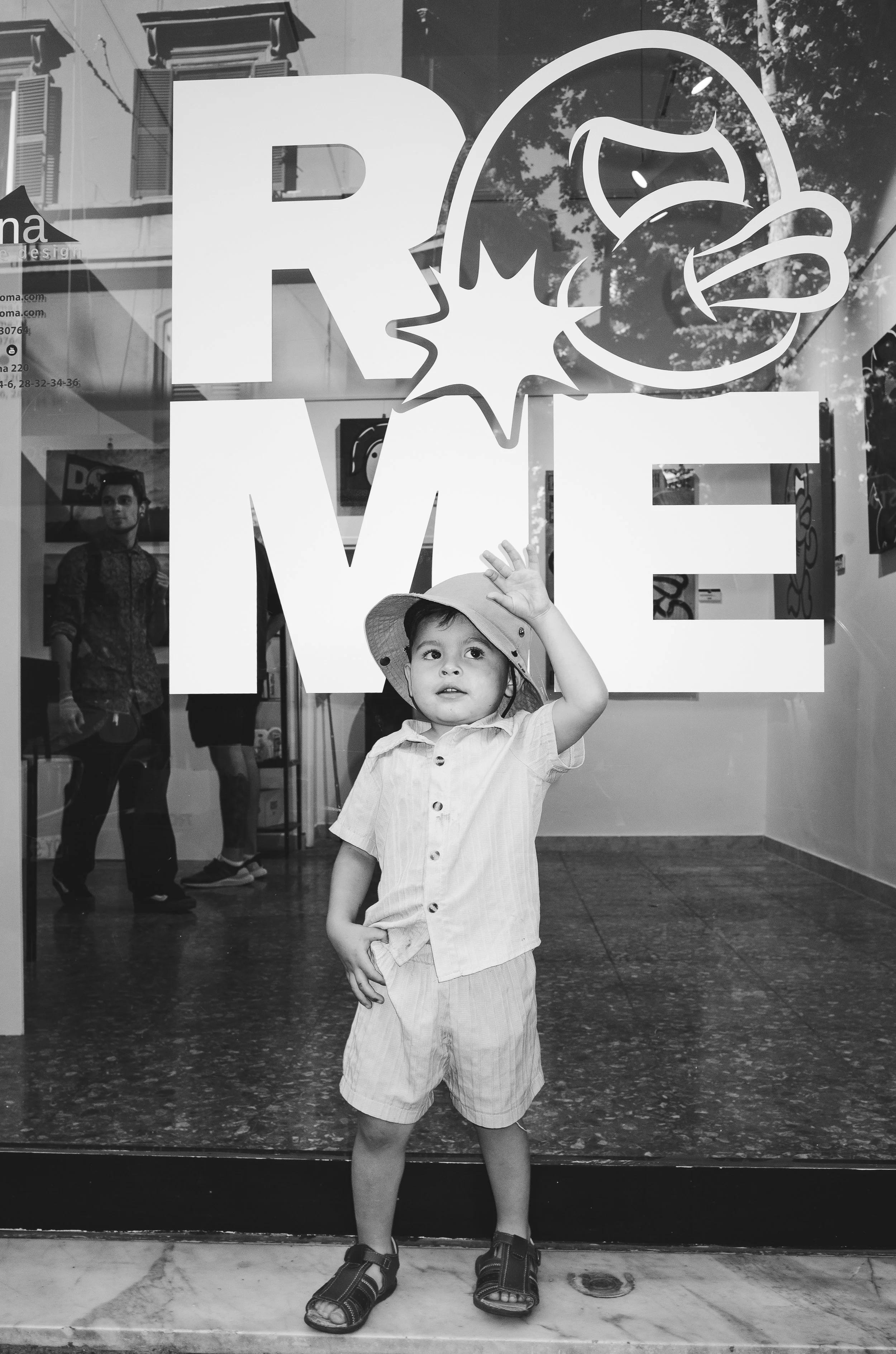 A young boy standing in front of a glass window with a 'LOVE' sign and a graphic of a bird's head. The boy is wearing a hat, a short-sleeved shirt, shorts, and sandals, and is waving to the camera.