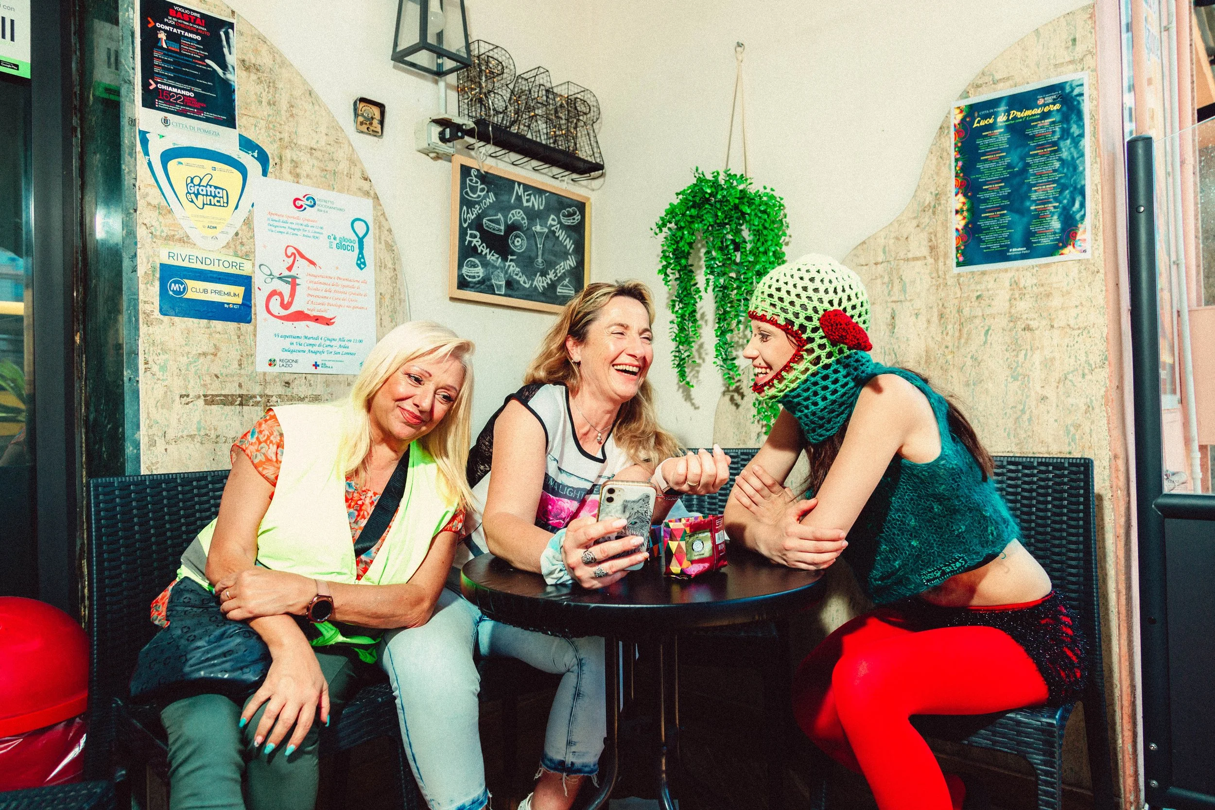 Three women sitting at a small black table in a casual restaurant and laughing. One woman is taking a photo or video with her phone. The woman on the right is wearing a green crochet hat with a red detail, a teal sleeveless top, and red stockings. Th