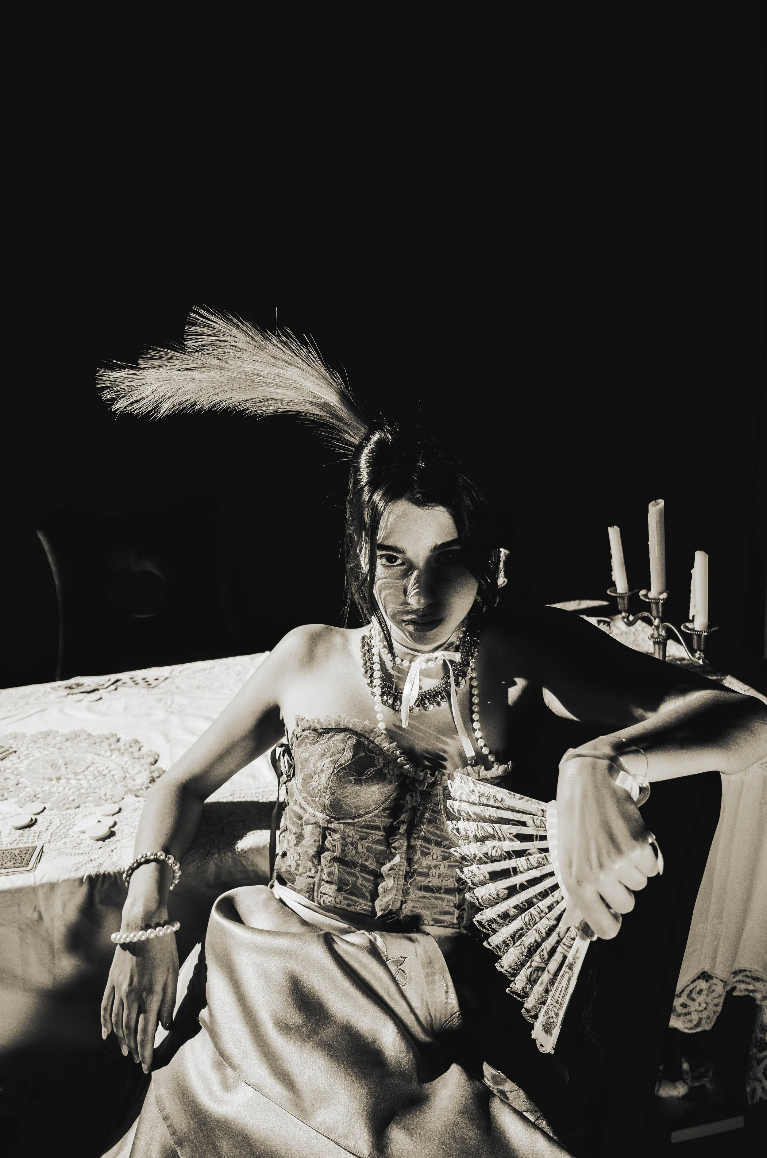 A woman dressed in vintage clothing with jewelry and a feathered headpiece sitting at a table with a lace tablecloth and candlesticks in the background, in a black-and-white photograph.