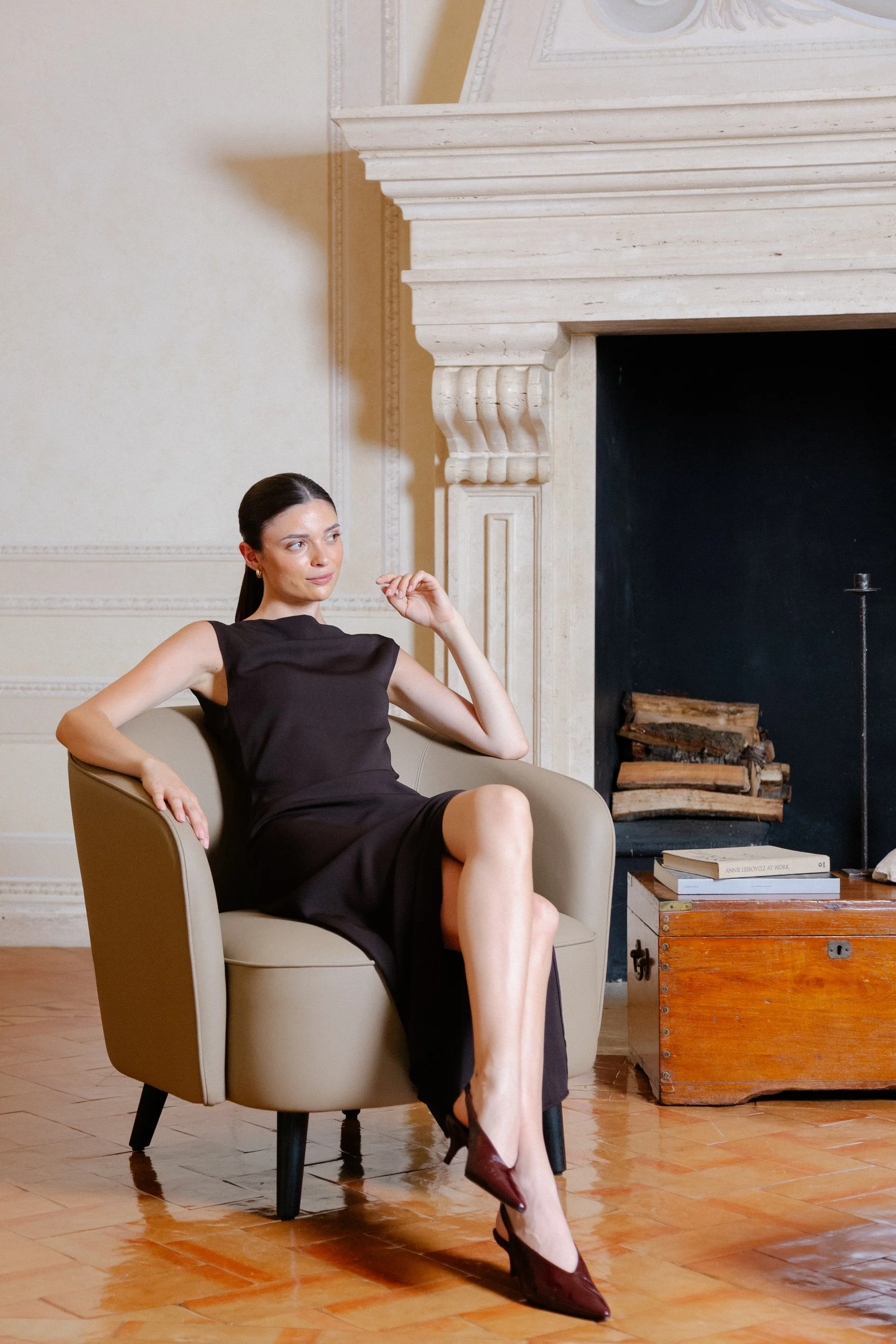 A woman with dark hair tied back, wearing a sleeveless black dress and maroon high heels, sitting on a beige armchair in front of a fireplace with stacked logs.