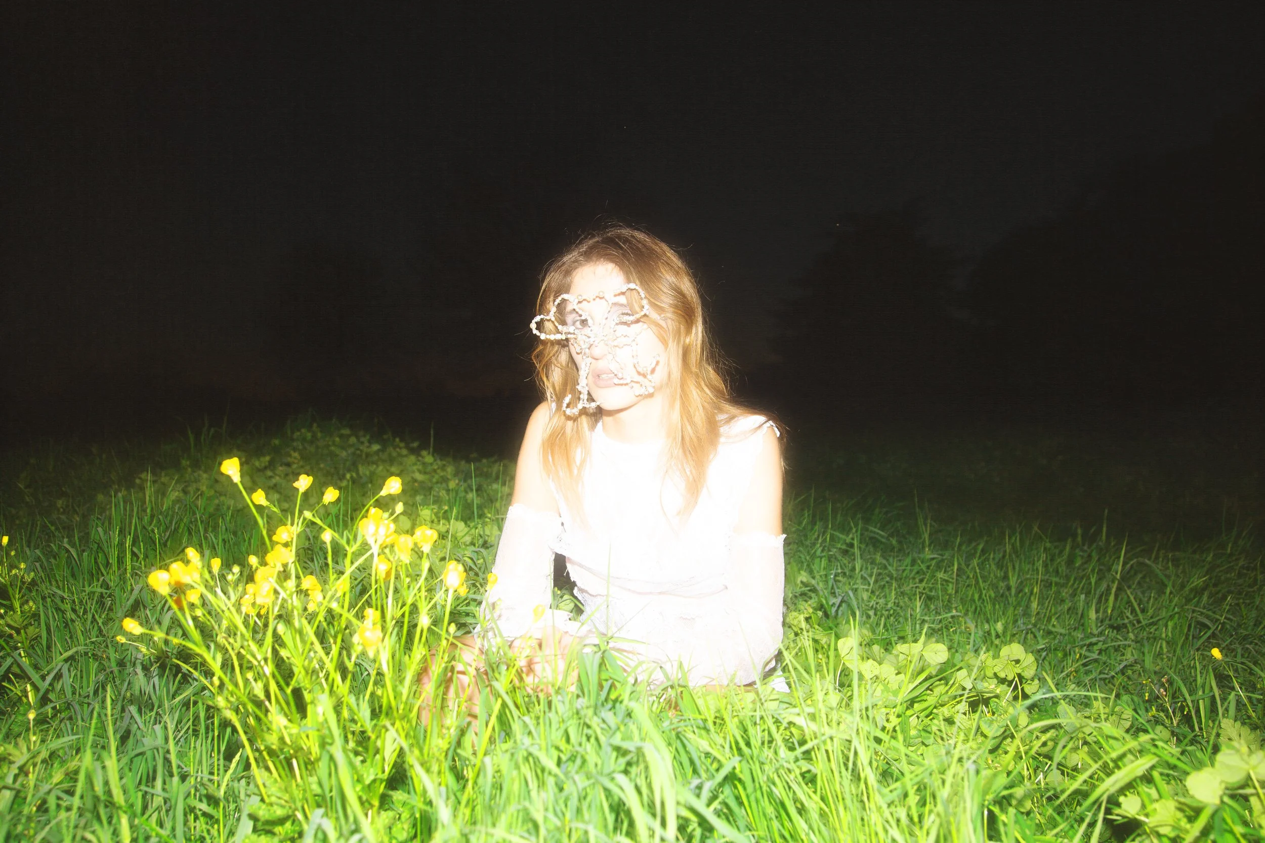 A person with light-colored hair sitting in a grassy field at night, illuminated by bright light, with a dark sky in the background. The person's face is obscured by a lace mask.
